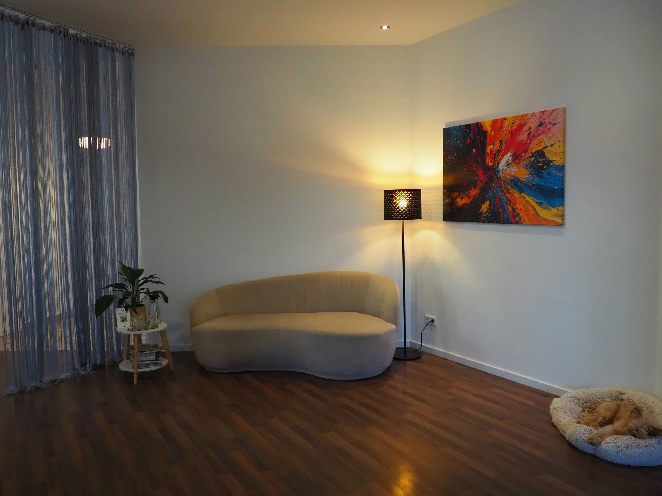 Gemütliches Wohnzimmer mit einer beige, modernen Couch, einem schwarzen Stehlampen, einem abstrakten Wandbild mit roten, blauen und orangefarbenen Farben, einem kleinen Beistelltisch mit Pflanzen und Zeitschriften, einer Tierbett mit schlafendem Hund