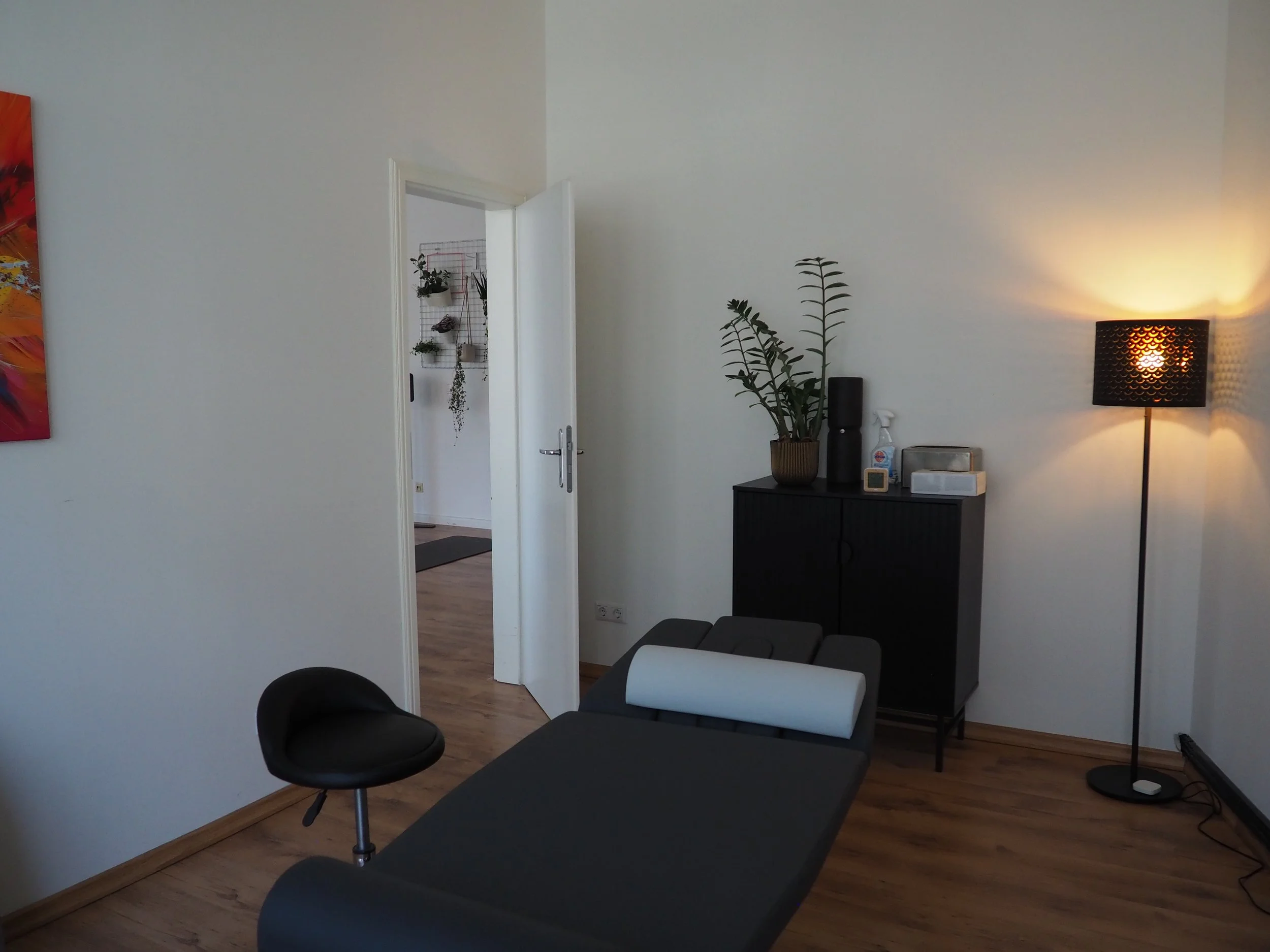 Behandlungsraum mit schwarzem Behandlungsliege, einer schwarzen Sitzhocker, einer schwarzen Kommode mit Pflanzen, einem digitalen Thermometer, einem Desinfektionsmittel und anderen Gegenständen, und einem Lampe mit warmem Licht.