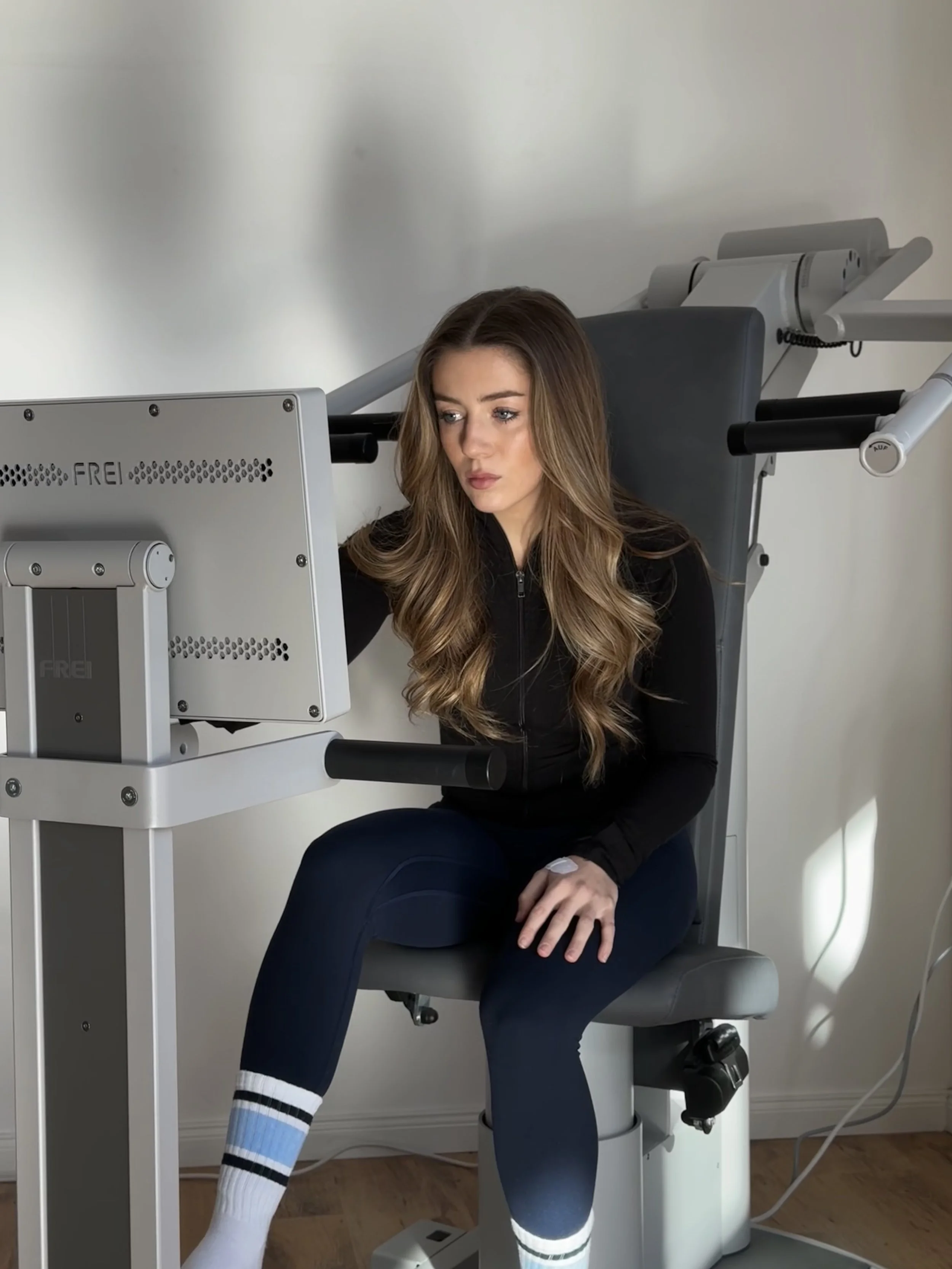 Junge Frau sitzt in einem medizinischen Gerät, das für physikalische Therapien verwendet wird, in einem Raum mit weißen Wänden.