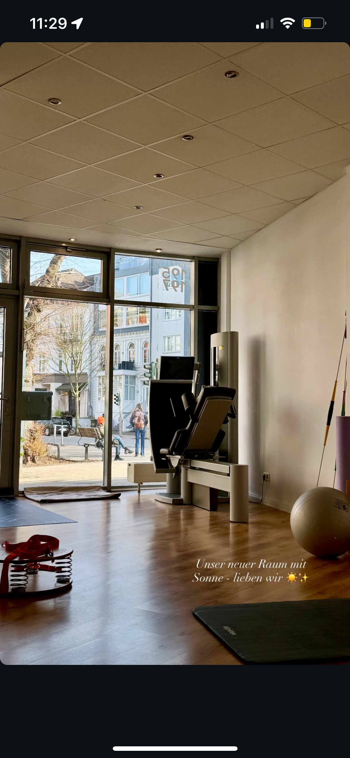 Ein Fitnessstudio mit großen Fensterscheiben, einem Trainingsgerät, Gymnastikmatten, einem Gymball und Fitnesszubehör. Es ist sonnig draußen, und es gibt eine Nachricht auf dem Bild: 'Unser neuer Raum mit Sonne - lieben wir'.