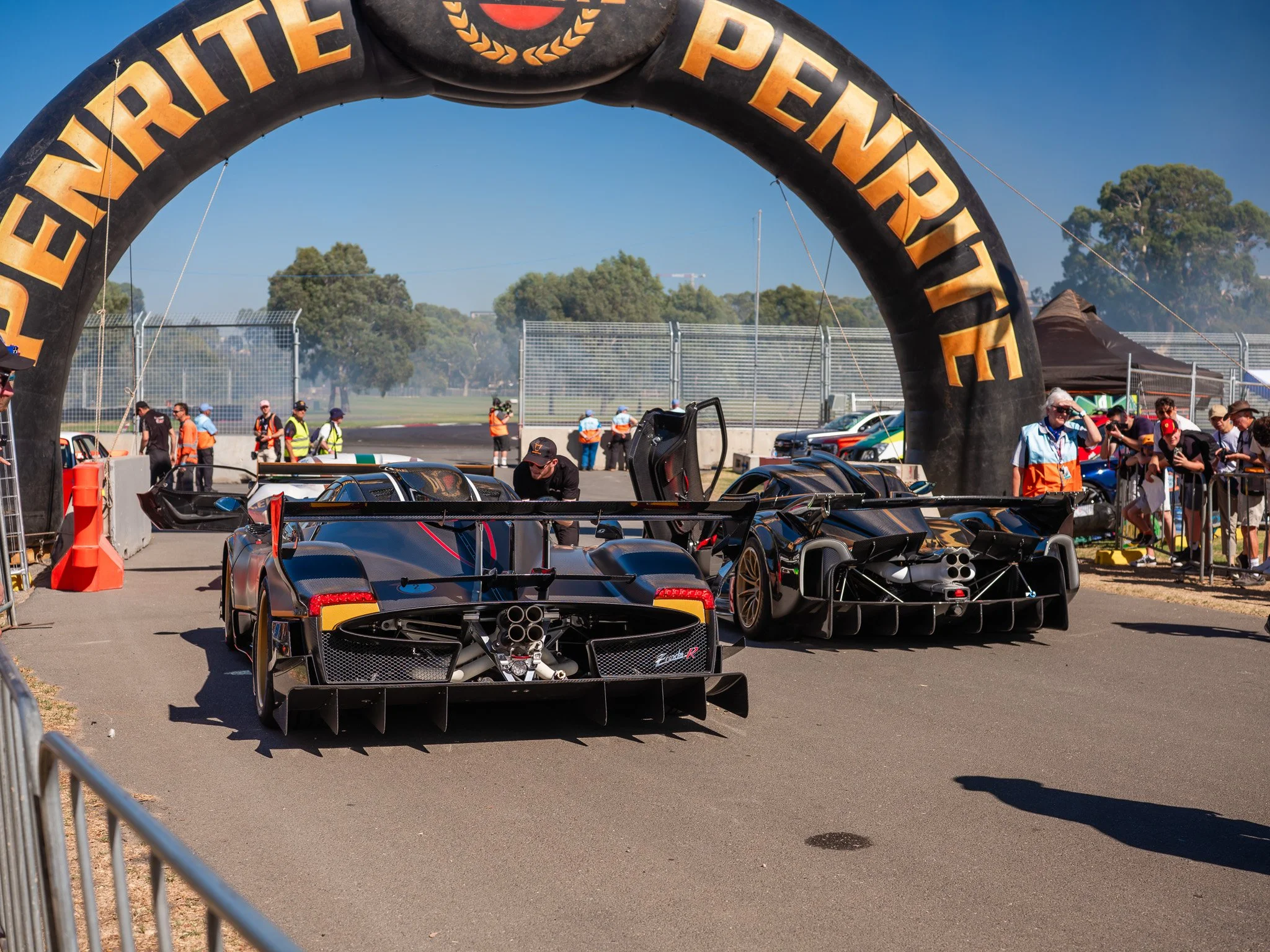 Two race cars parked under an inflatable archway reading 'PENTRITE'. Behind them are race officials and spectators, with some smoke in the background and trees in the distance on a sunny day at a racing event.