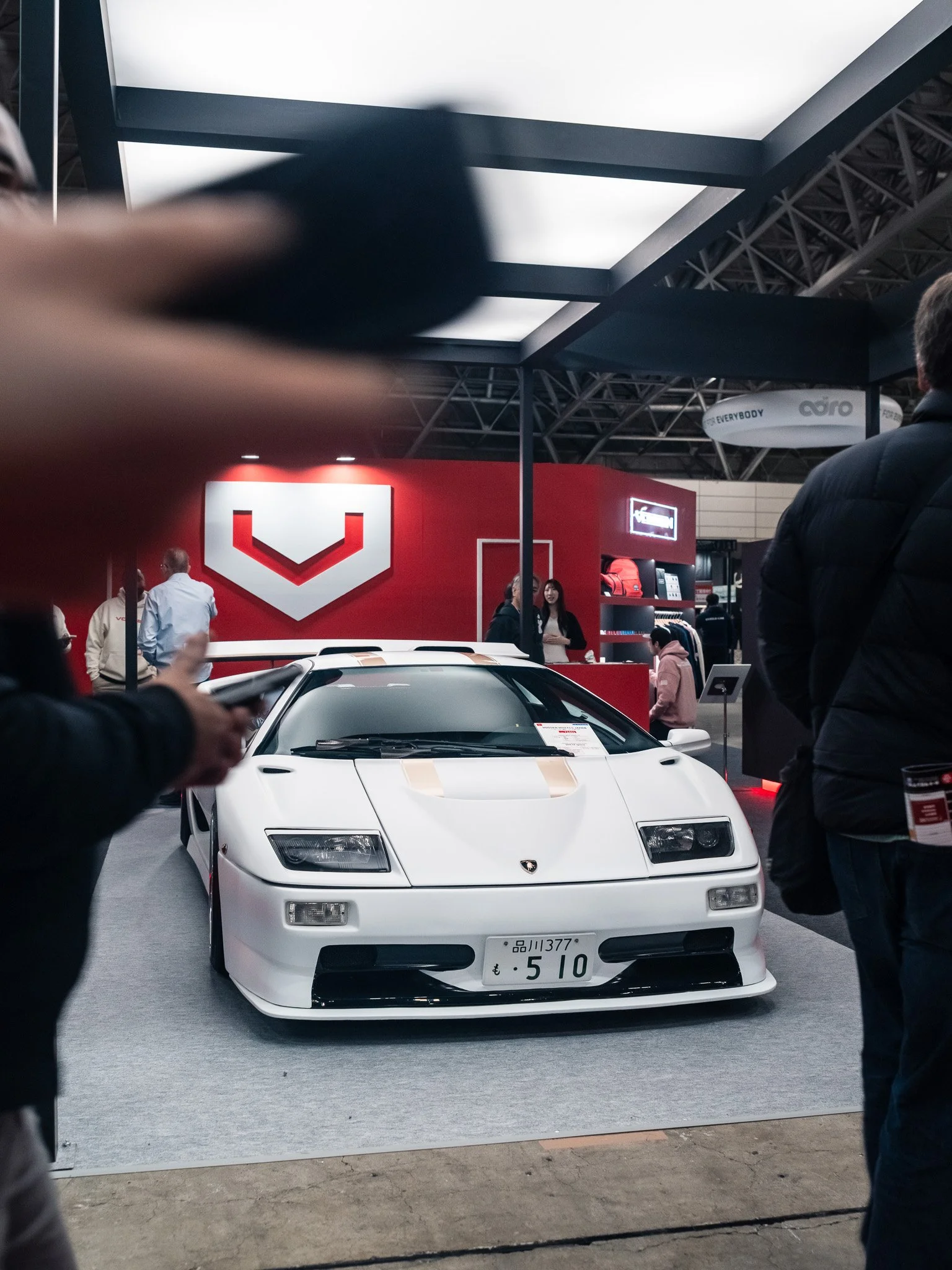 A white sports car on display at an auto show with a red backdrop featuring a logo. People are gathered around, some taking pictures, inside a large exhibition hall.