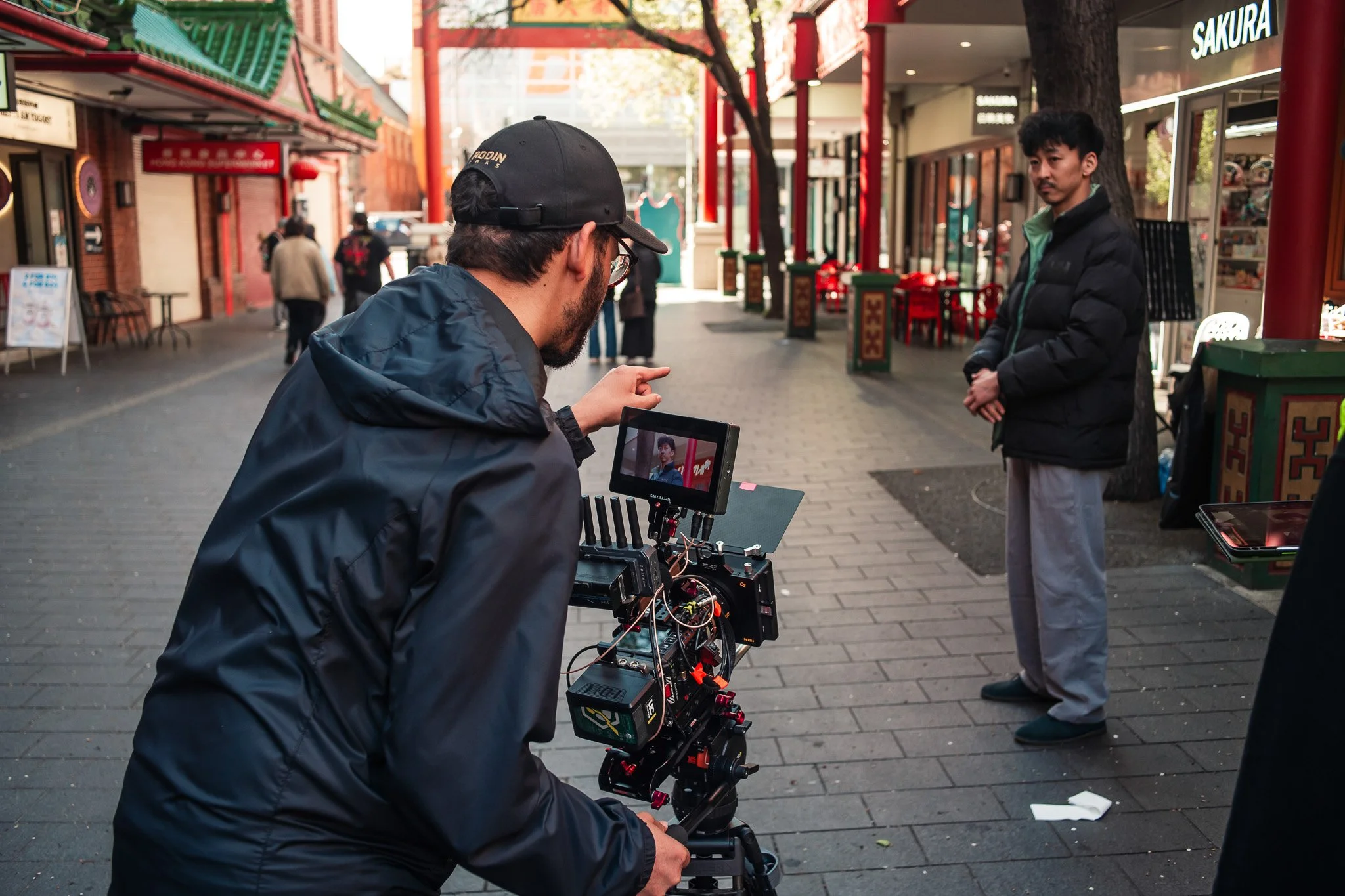 Filmmaker operating camera on busy city street with pedestrians and storefronts in background.