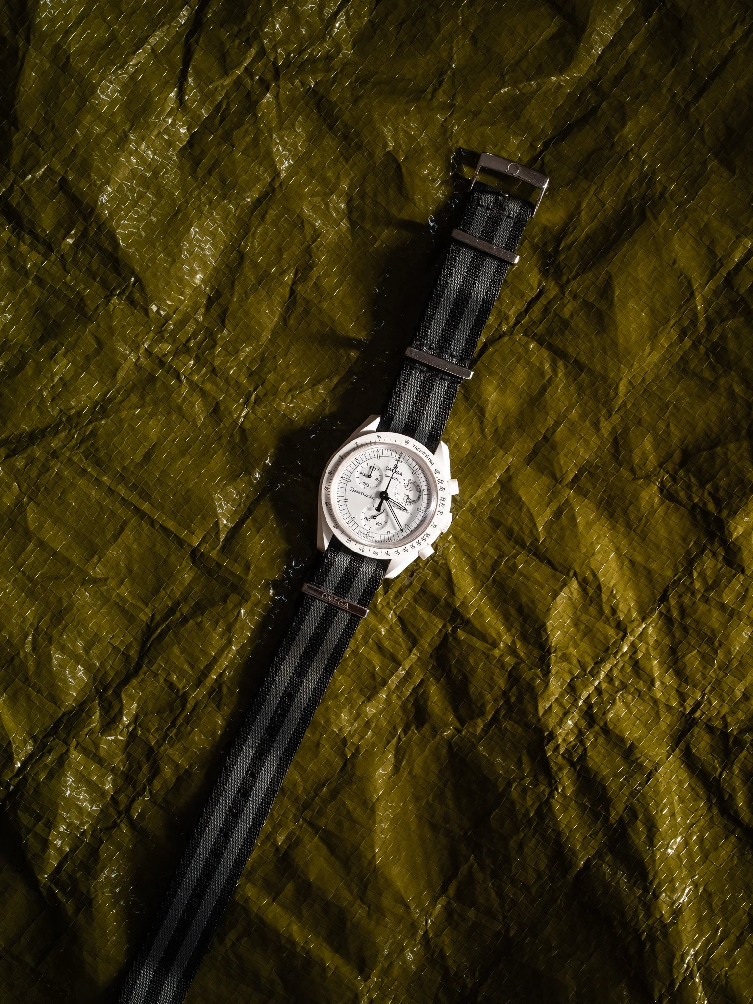 A white chronograph wristwatch with a black and gray striped fabric strap on a crinkled olive green surface.
