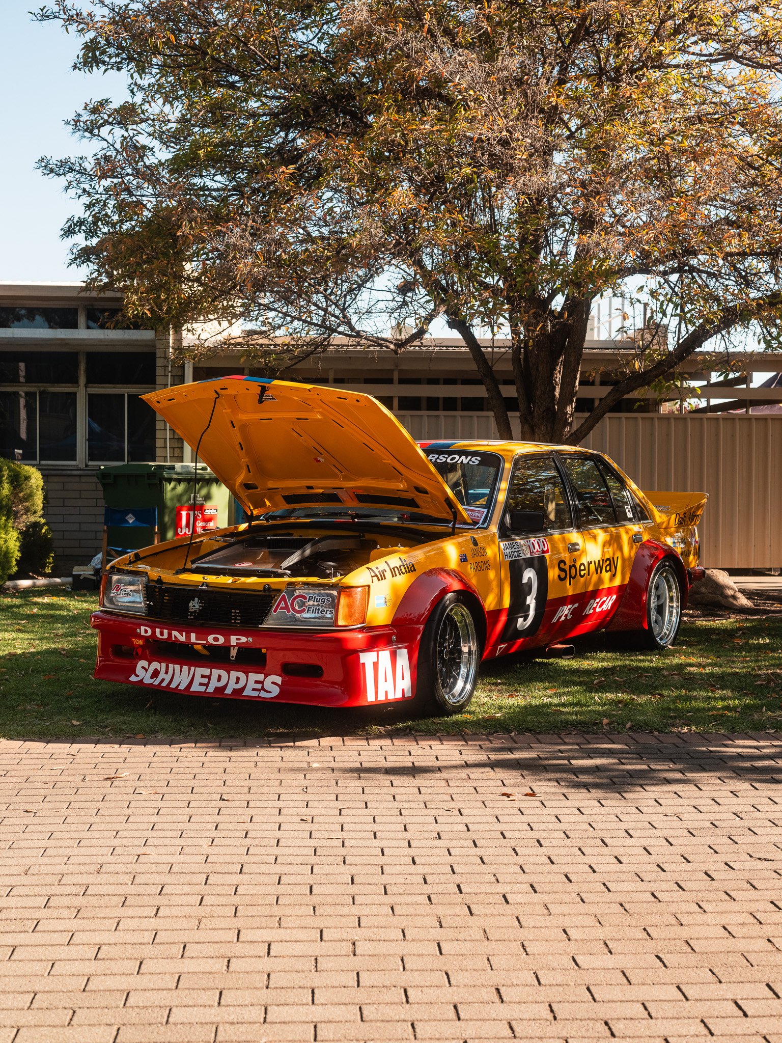 A classic racing car with yellow and red livery, open hood, parked on grass and brick pavement, with a tree and building in the background.