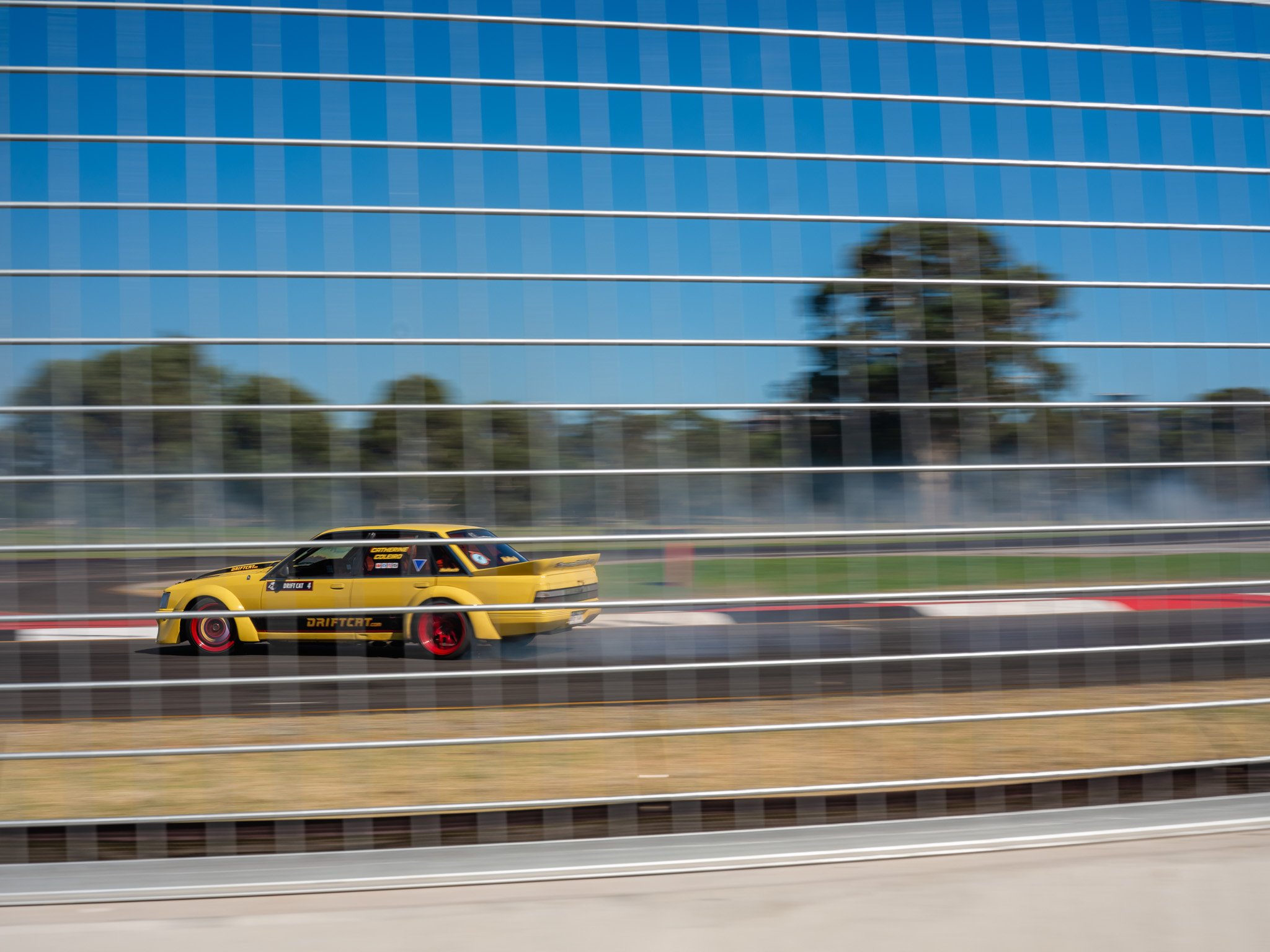 A yellow drift car with red wheels driving at high speed on a race track, seen through a glass barrier with a reflection of trees and a blue sky.