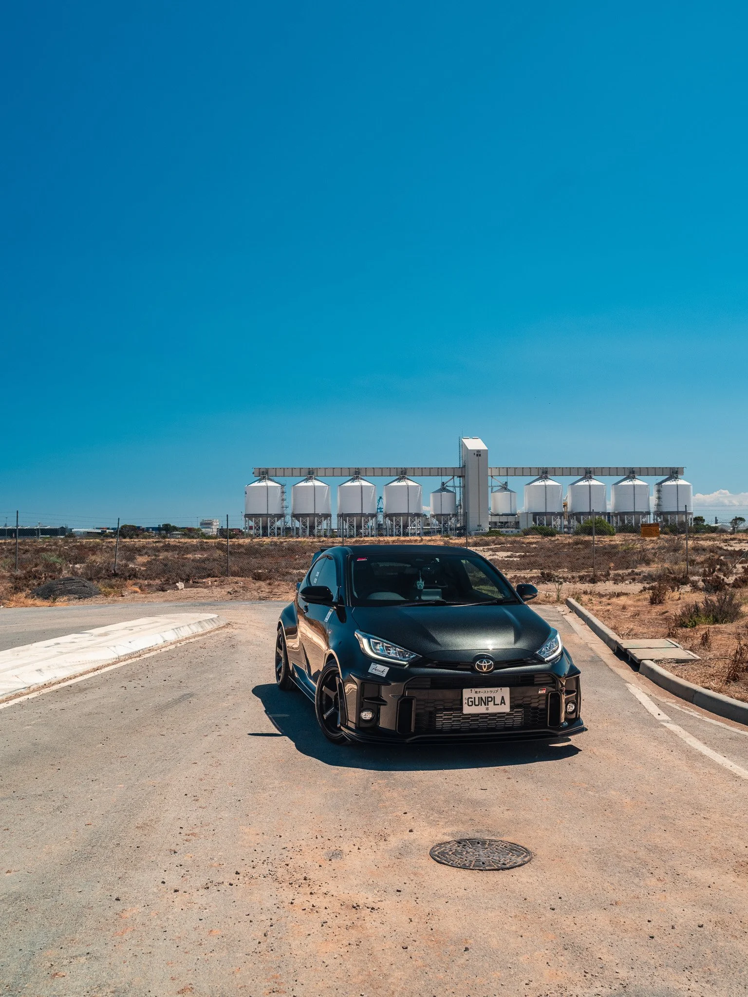 A black Toyota sports car with a custom front bumper, lowered stance, and a license plate that reads 'GUNPLA,' parked on an empty road in a dry, desert-like area with industrial silos in the background under a clear blue sky.