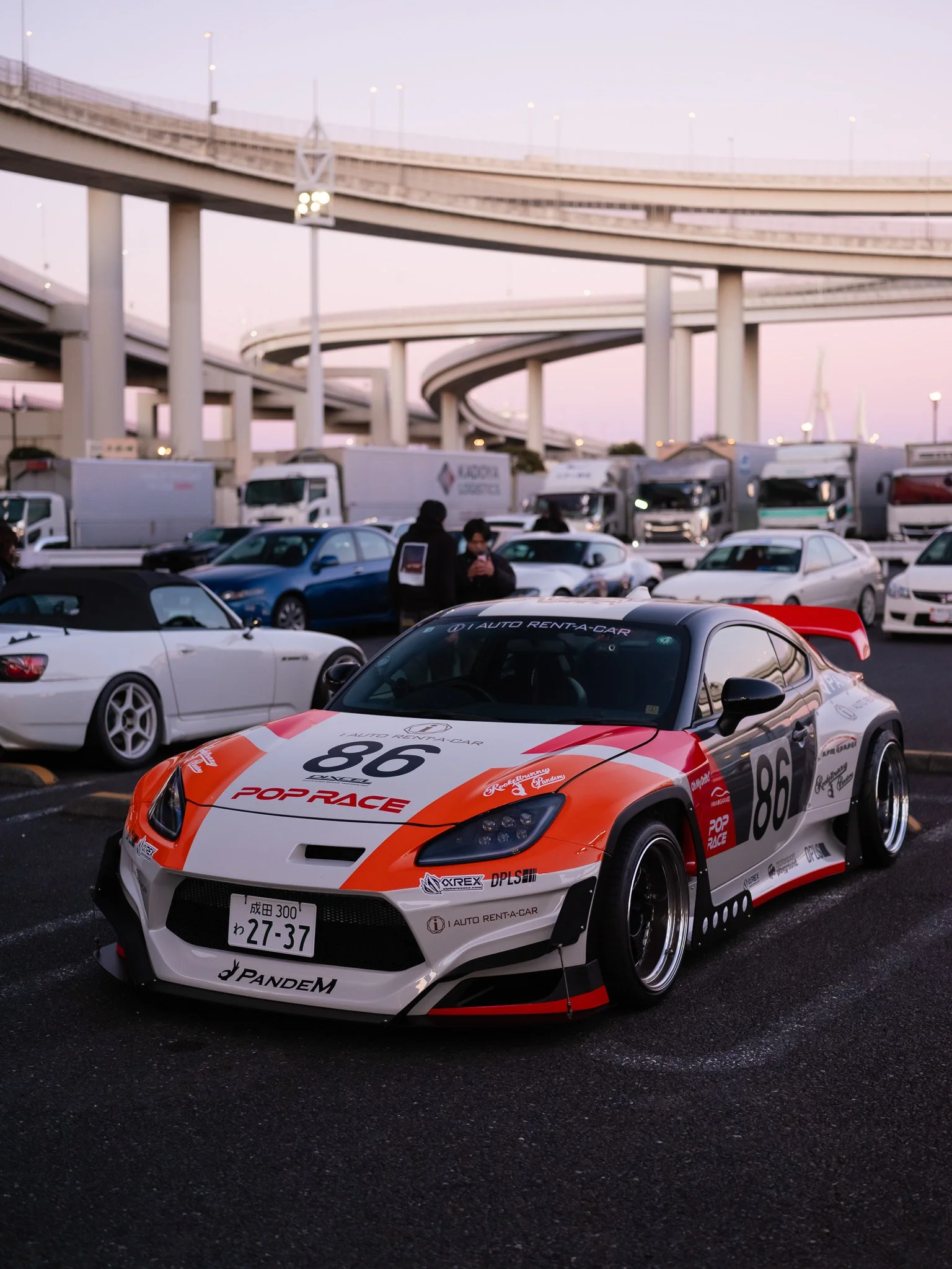 A race car with racing decals and the number 86 parked in a parking lot with other cars and trucks, under an overpass at sunset.