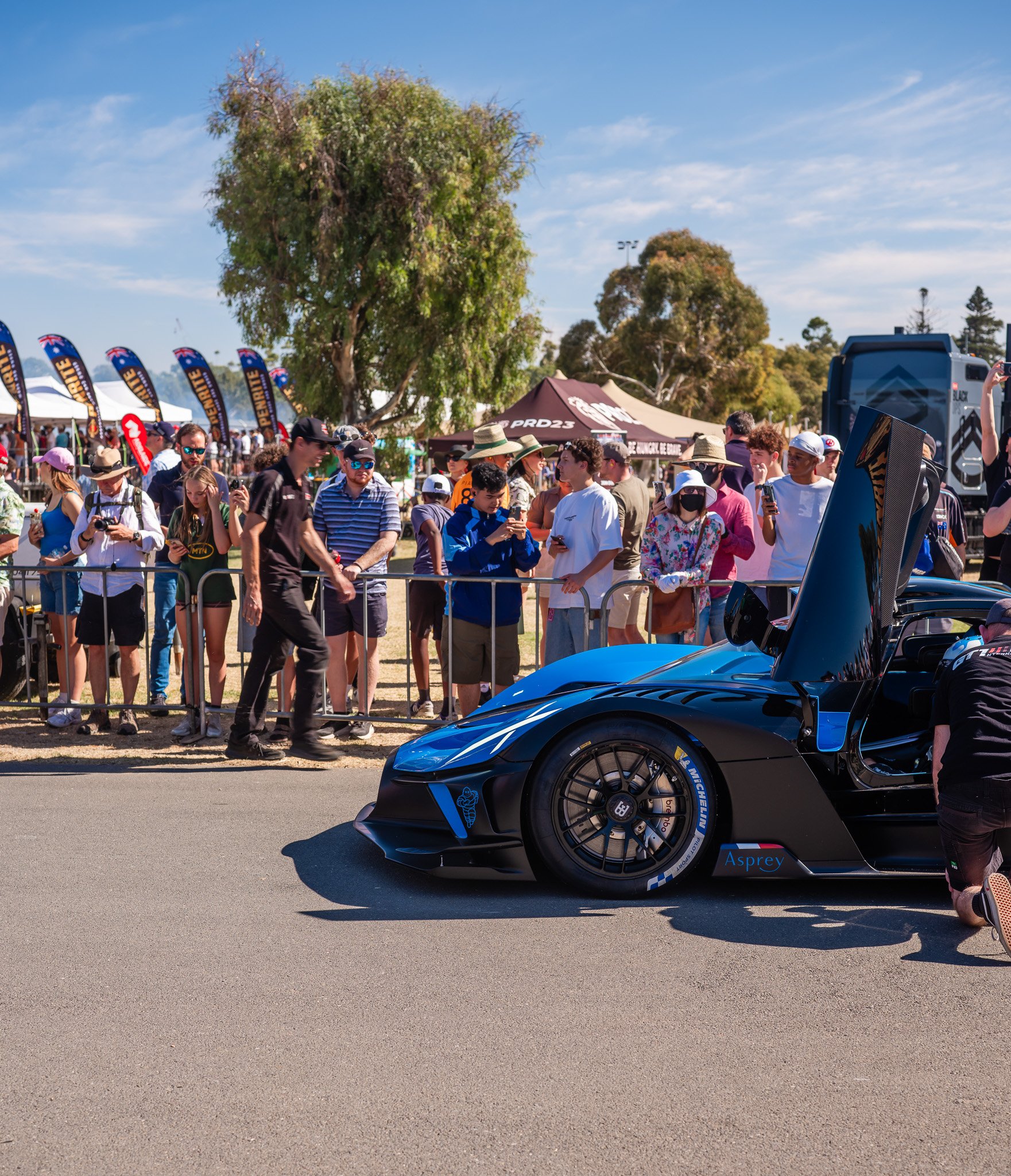 People attending a car show look at a sleek blue sports car with its door open. The crowd is behind a barrier, many taking photos and videos, with tents and banners in the background under a clear blue sky.