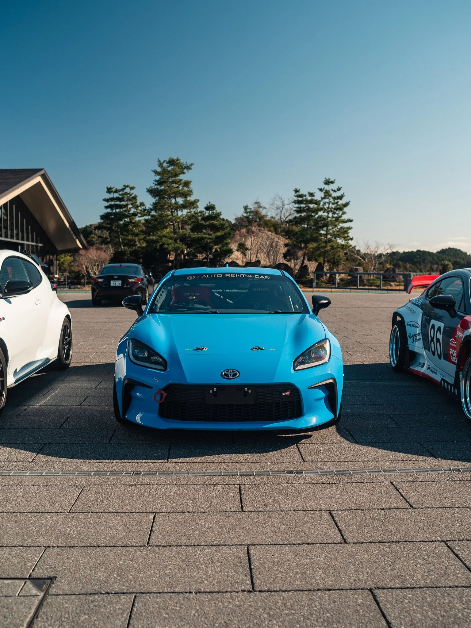 A blue Toyota racing car parked in a lot beside other cars, with trees and a building in the background, under a clear blue sky.