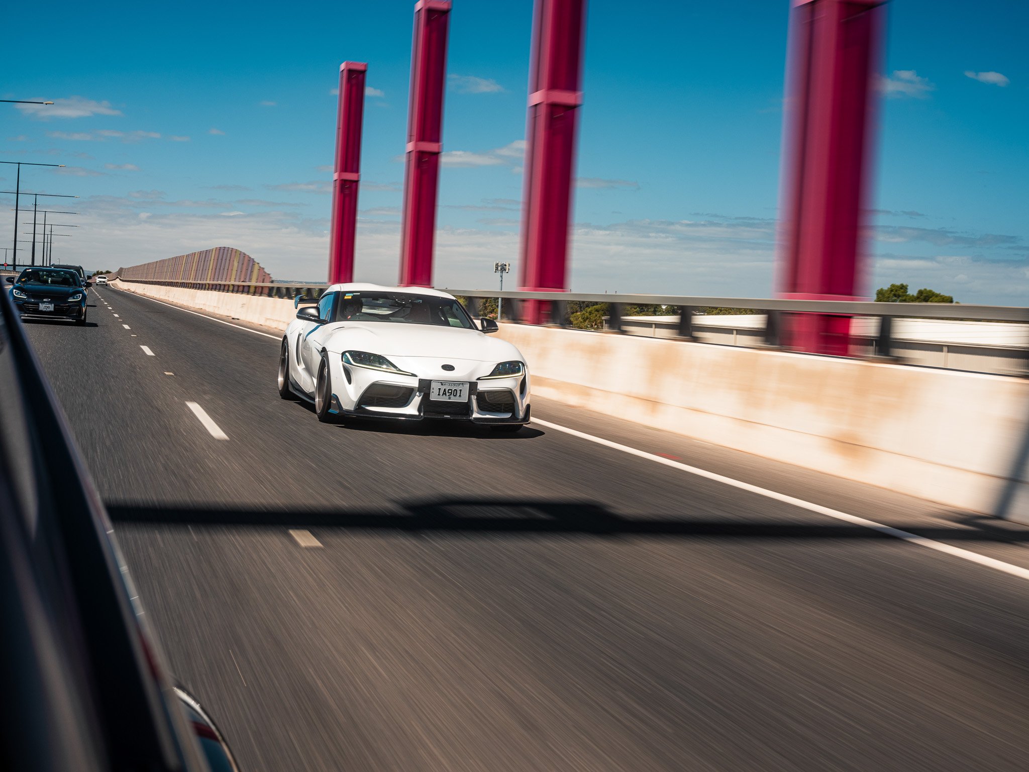 A white sports car driving on a highway with a bridge's red support beams on the right side under a blue sky with some clouds.