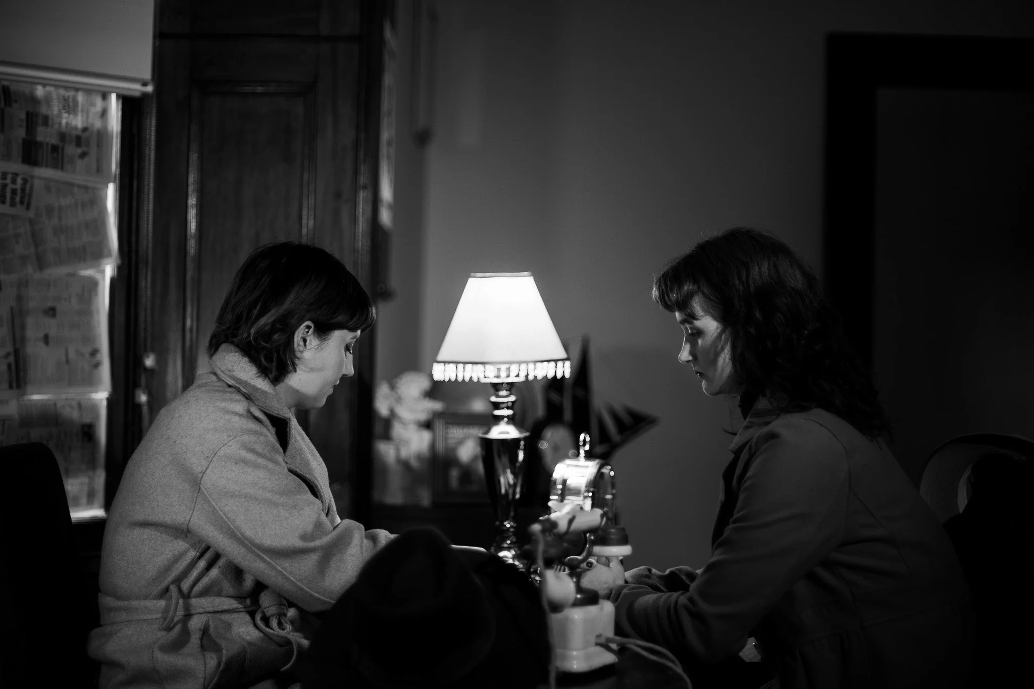 Two women sit at a table with a lamp between them in a dimly lit room, engaging in a serious or intimate conversation.