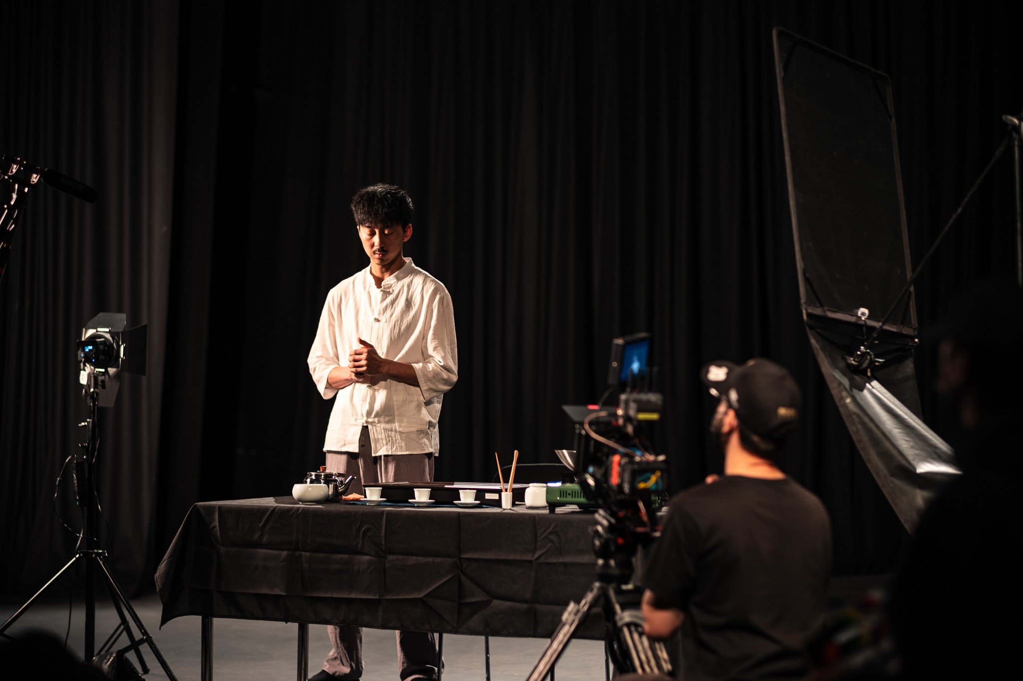 A man in traditional white clothing stands with hands clasped in front of a black curtain on a stage, surrounded by filming equipment including cameras and lighting, with a table setup in front of him.