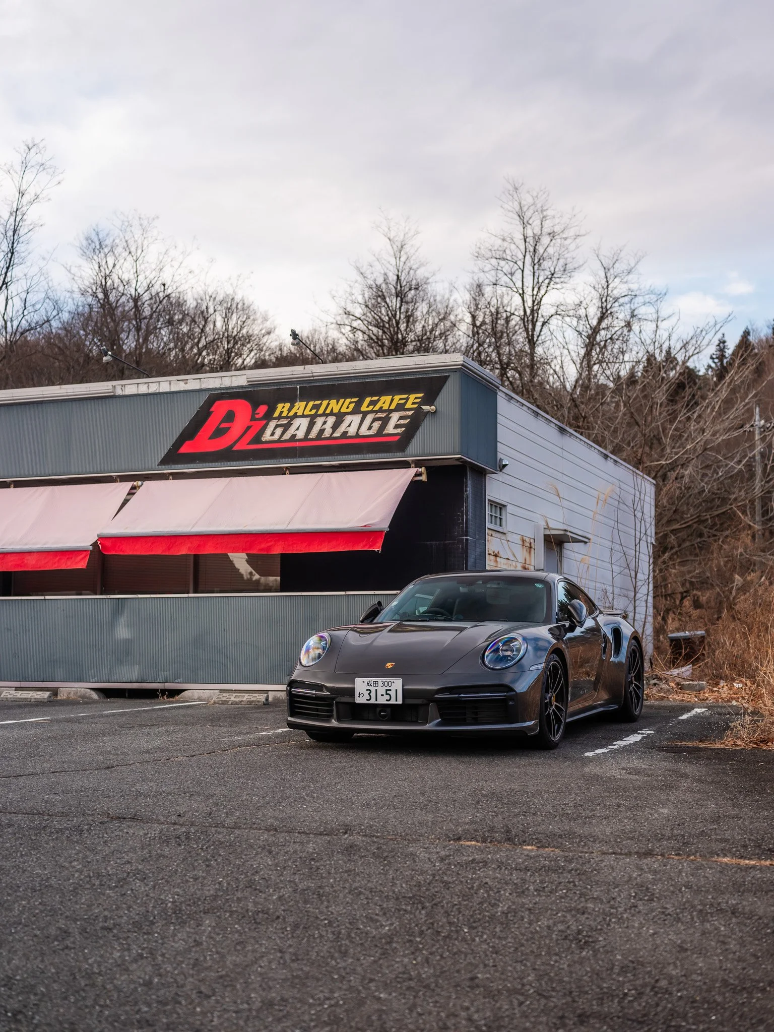 A black Porsche sports car parked in front of a building with a sign that reads 'D1 Racing Cafe Garage' under a cloudy sky, with leafless trees in the background.