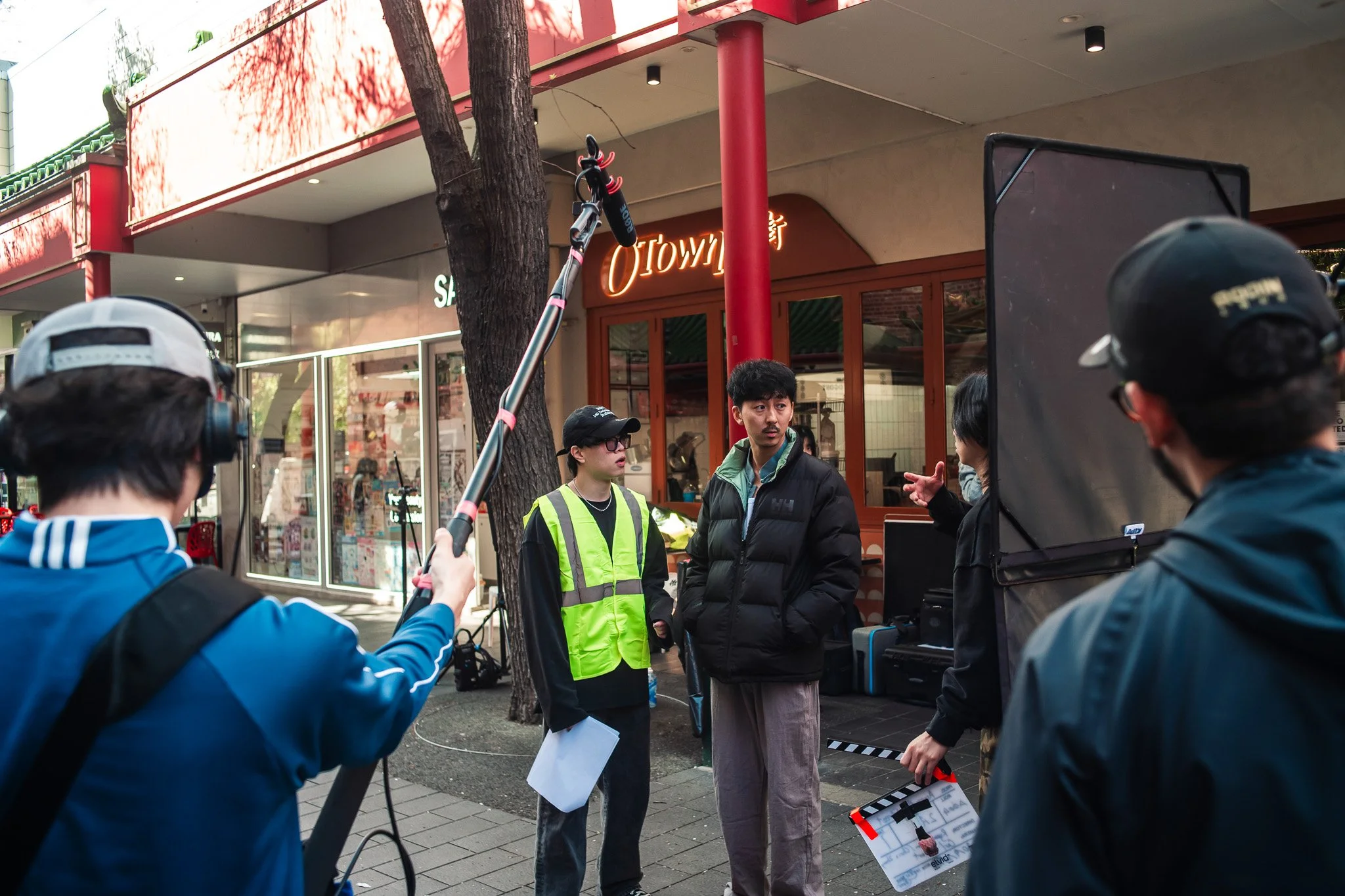 Filmmaking crew setting up camera and lights on a city sidewalk while two actors prepare for a scene outside a storefront with a red and beige facade.