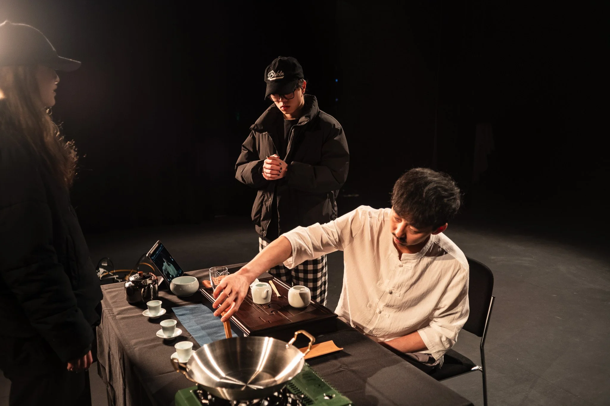 A man in a white shirt is sitting at a table with tea cups, a teapot, and a camera, pouring tea while two women, one in a black hat, watch.