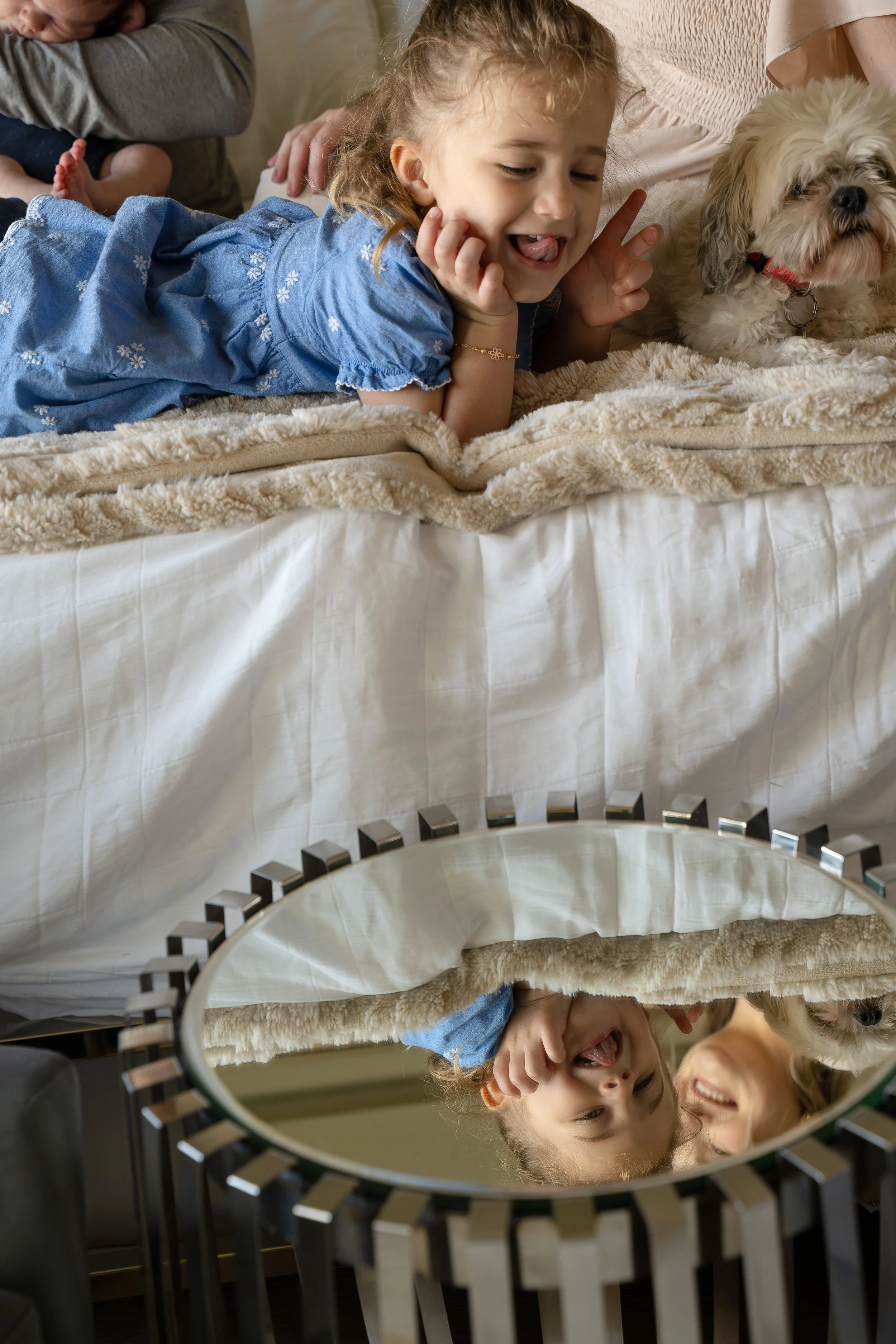 A young girl with curly hair lying on a bed with a fluffy blanket, making a silly face with her tongue out, in front of a mirror that reflects her face and a woman smiling nearby.  The Woodlands & Houston Heights In Home Newborn & Family photographer