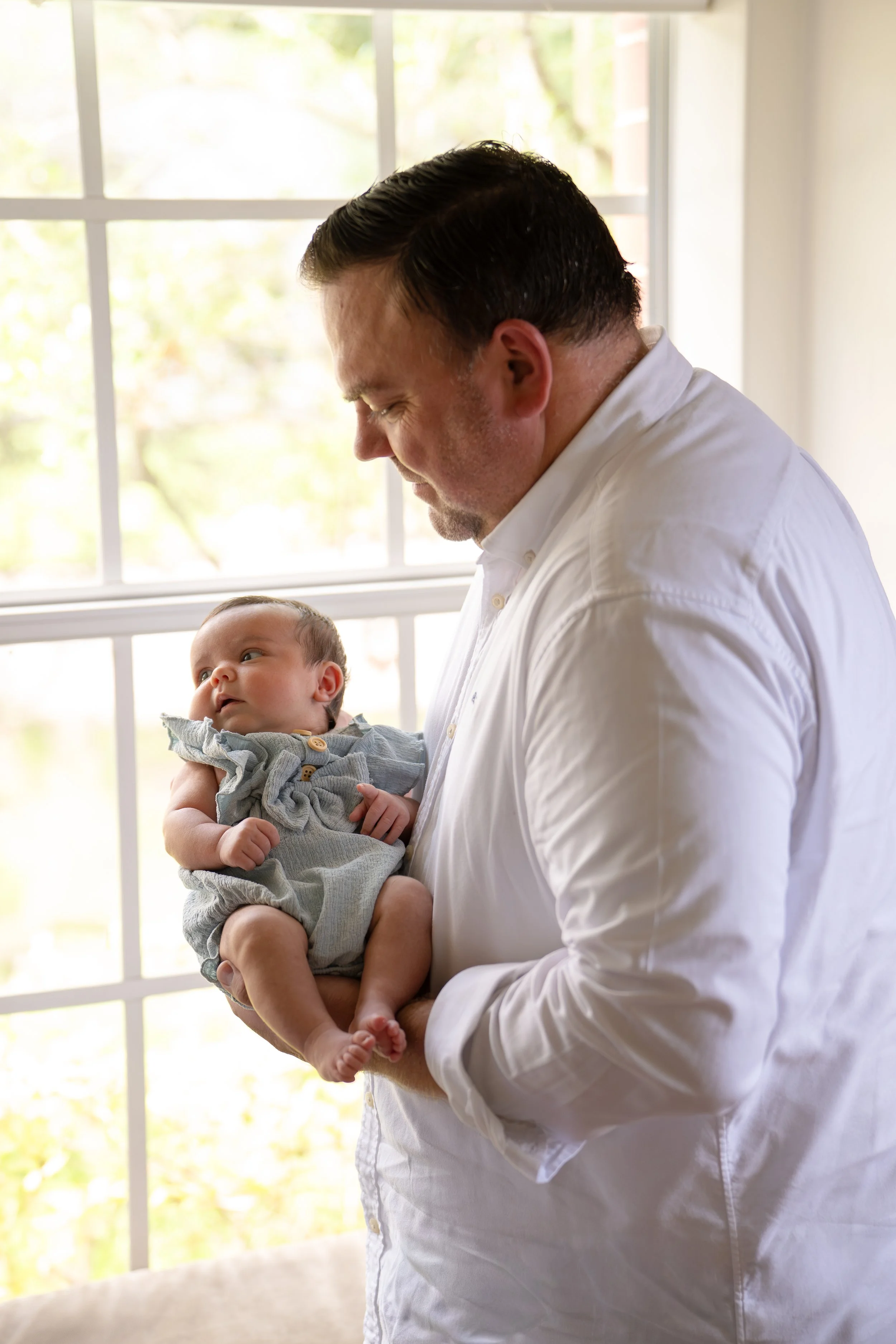 A man with dark, slicked-back hair in a white shirt is holding a baby with light brown hair, gray clothing, and a curious expression, standing in front of a window with a view of trees outside. in the home of his The Woodlands home.