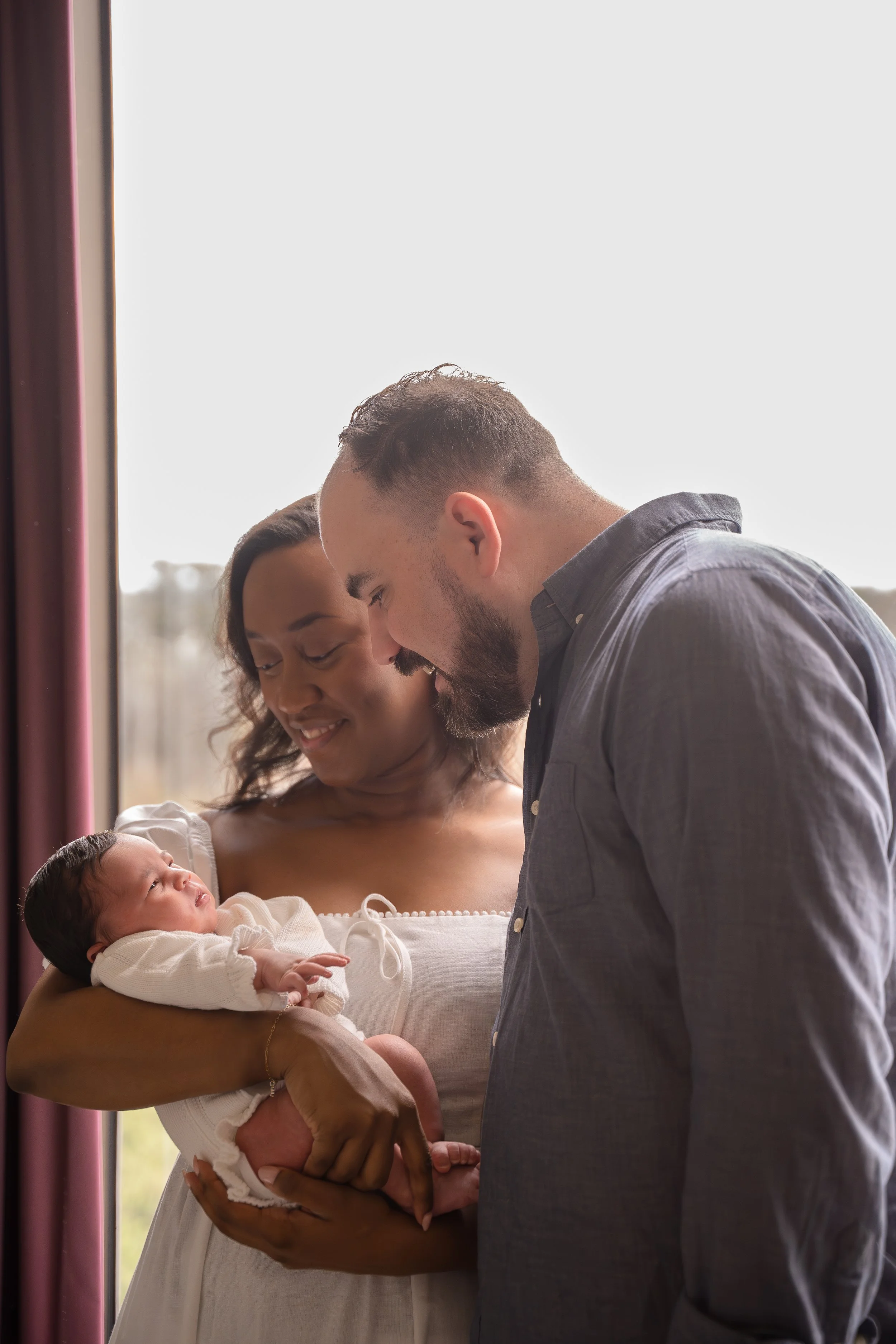 A woman holding a newborn baby, with a man leaning over them, in a room with a large window and natural light in the Houston Ruby Postnatal. 