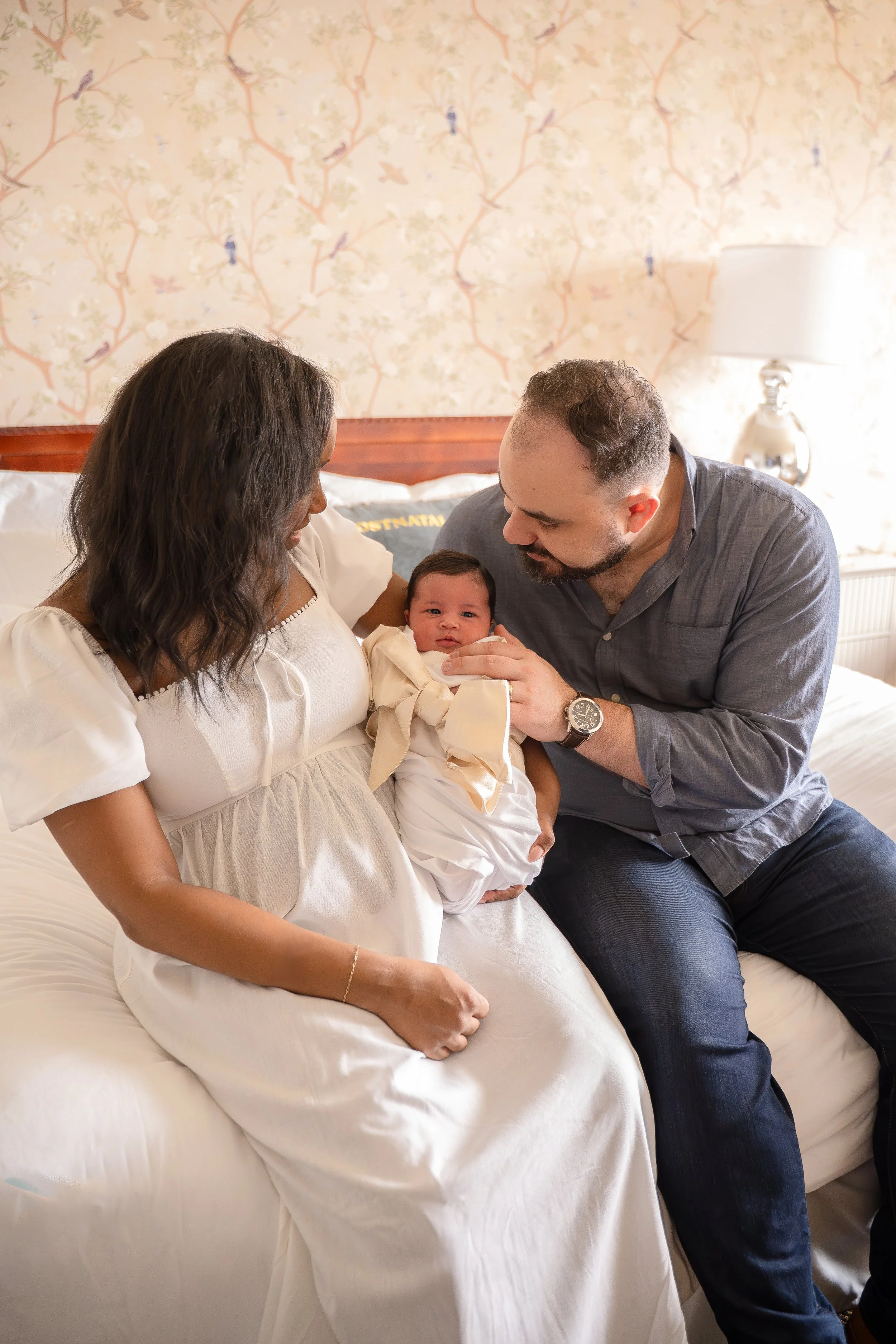 A woman and a man are sitting on a bed, holding their baby together. The woman is wearing a white dress and the man is wearing a gray shirt and jeans. The baby is wrapped in a blanket and looking at the camera, in the Houston Ruby Postnatal. 