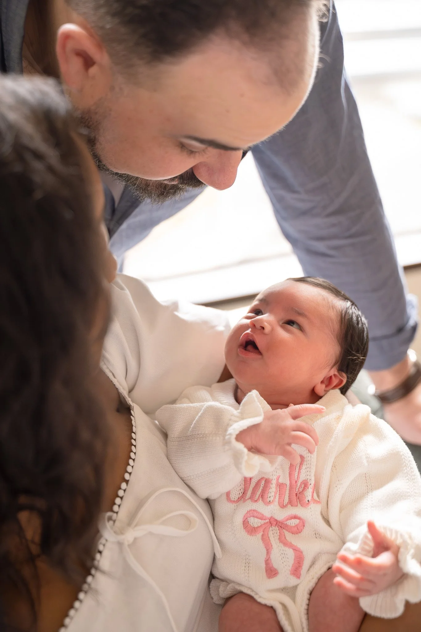 A man is leaning in close to a baby girl, holding her gently as she looks up at him with a smile, in the Houston Ruby Postnatal. 