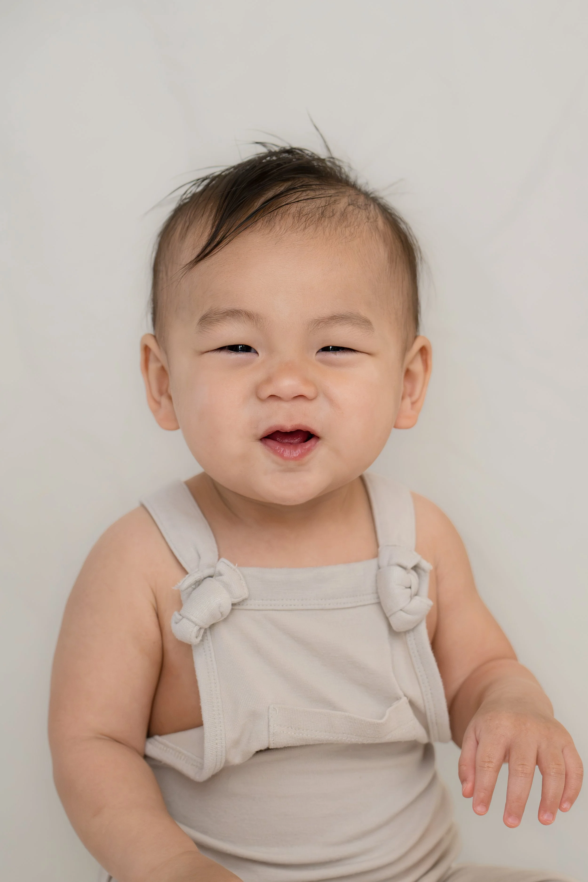 Close-up of a smiling Asian baby with dark hair, wearing a beige sleeveless top with knot details on the shoulders, against a plain white background, in Katy, TX at home.