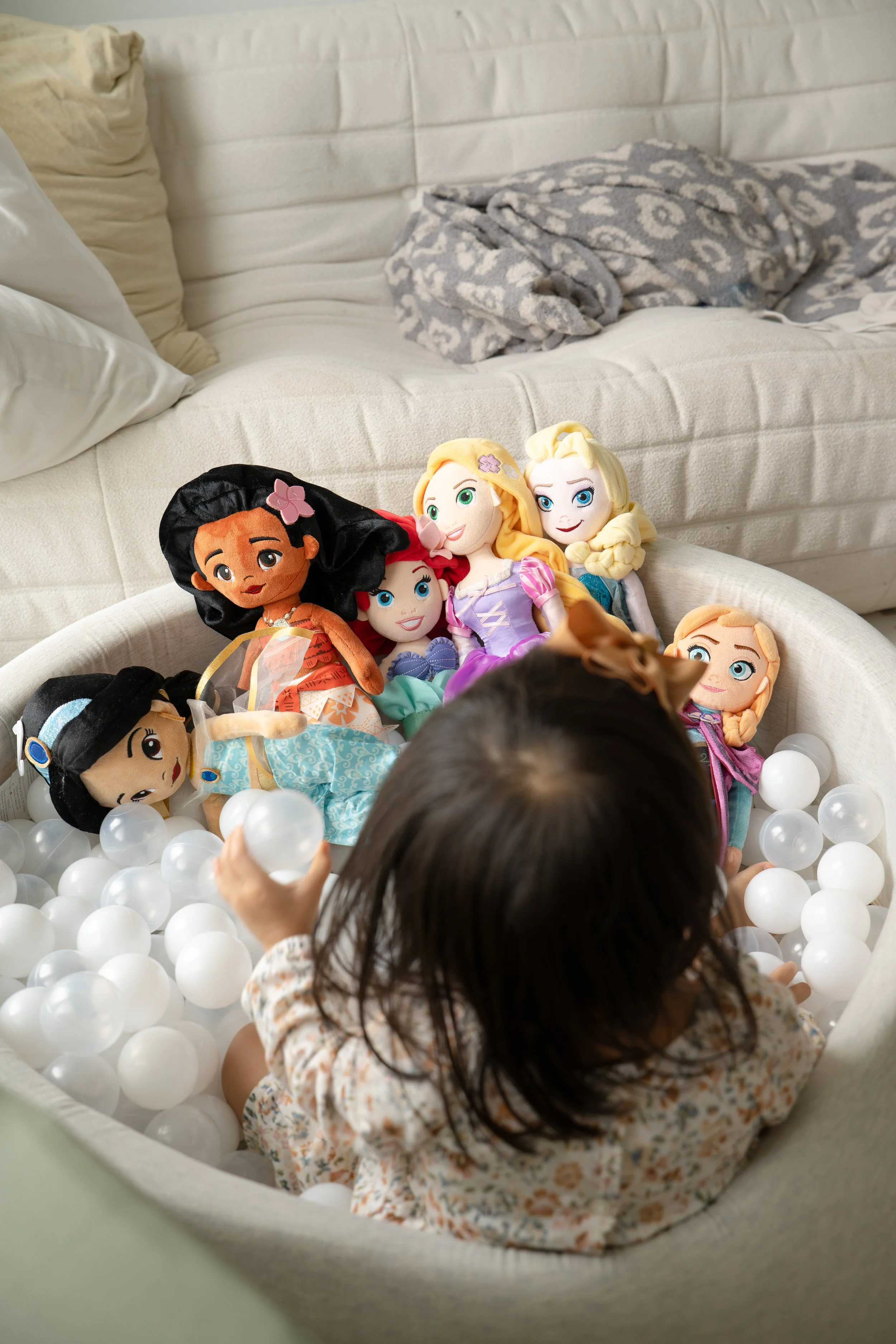 A young girl with dark hair playing in a ball pit filled with white plastic balls, surrounded by plush Disney princess dolls including Rapunzel, Moana, and Elsa, in a cozy room at home in Katy, TX.