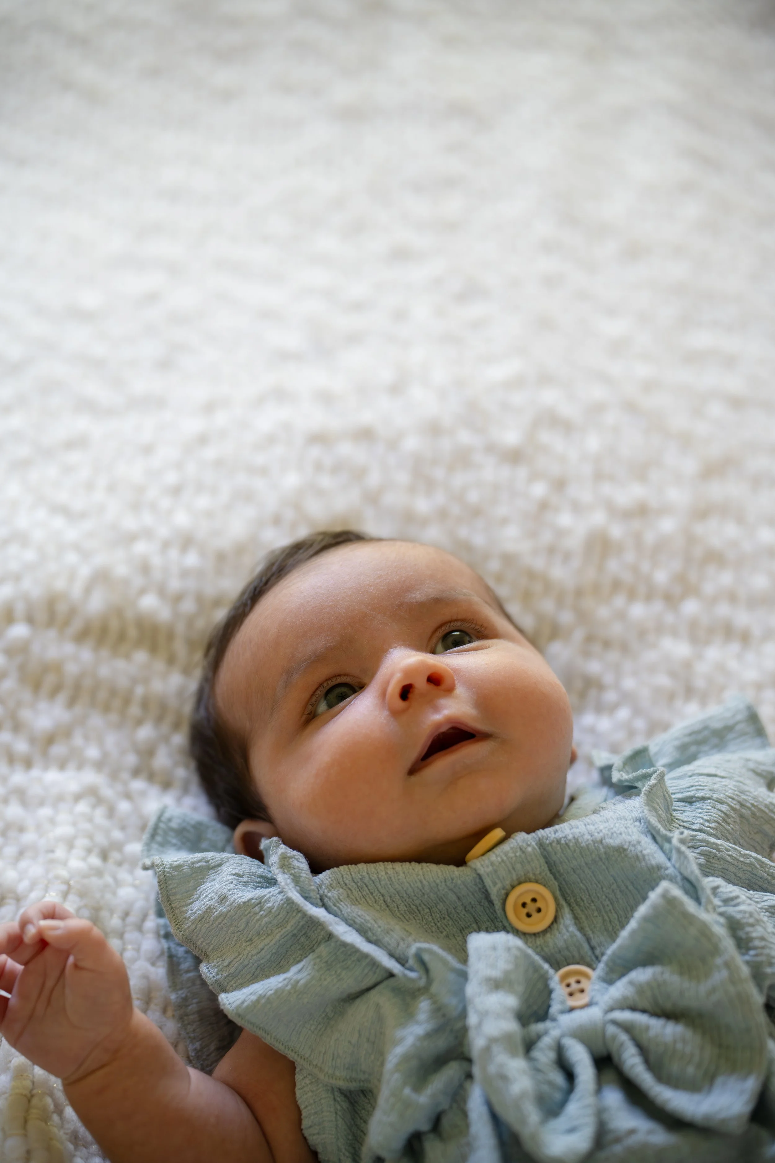 Close-up of a baby lying on a soft, cream-colored textured blanket, with a slight smile and wide eyes, wearing a light green outfit with yellow buttons. The baby is laying on the bed of their The Woodlands home.