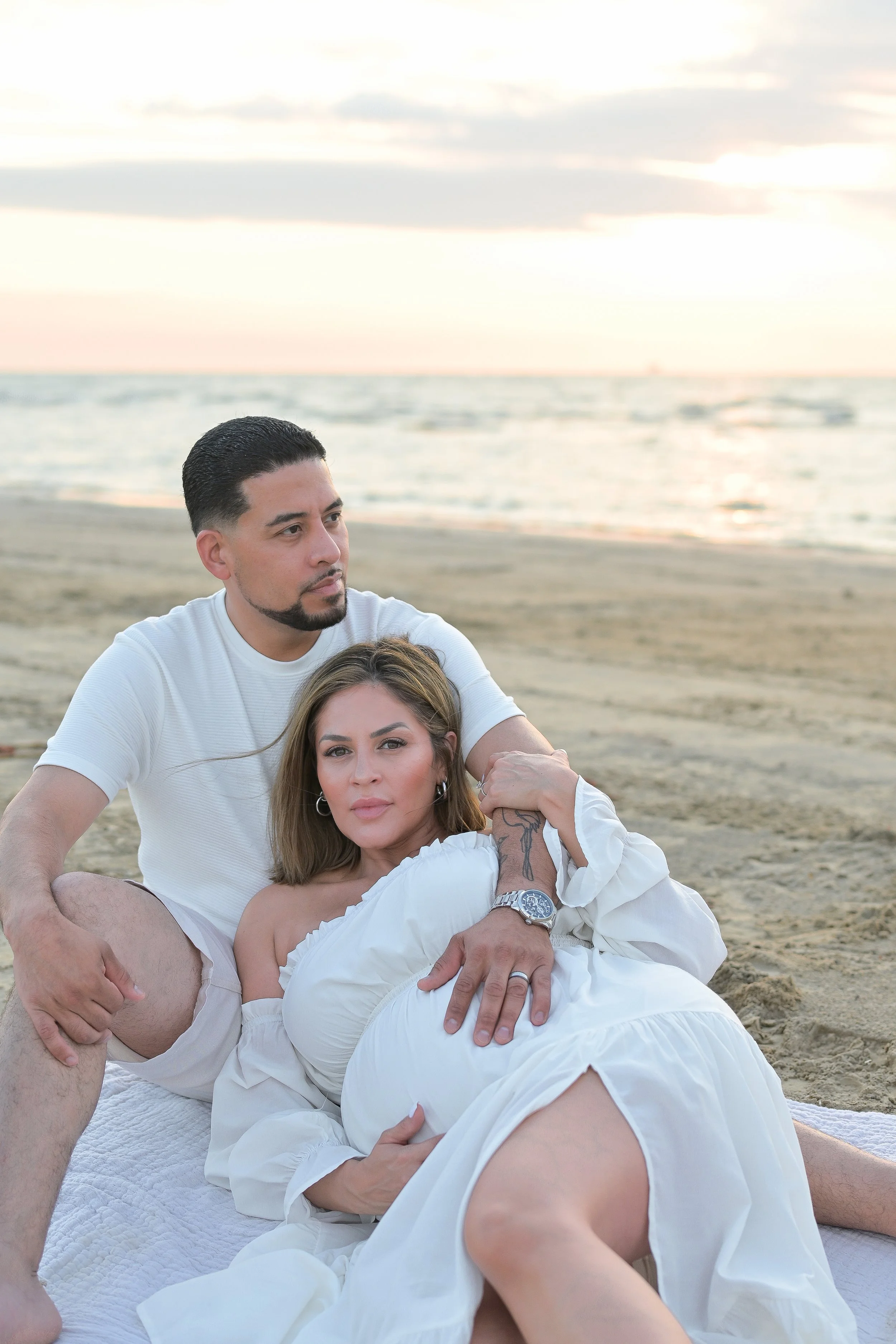 houston couple sitting on the beach at sunrise with maternity belly showing