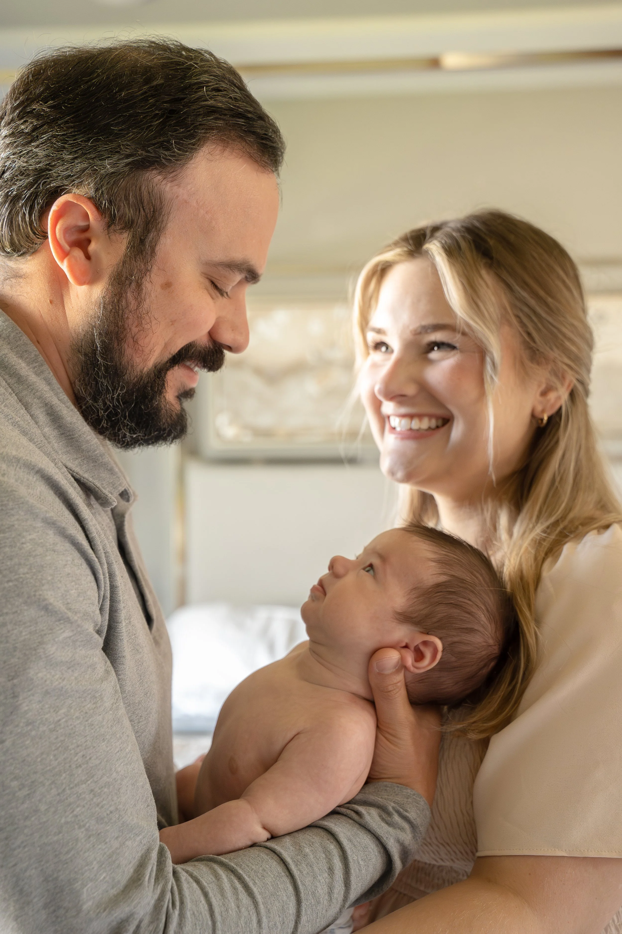 A man with a beard and a woman with blonde hair smiling at each other as they hold a newborn baby in a hospital room.  The Woodlands & Houston Heights In Home Newborn & Family photographer providing luxury artwork albums.