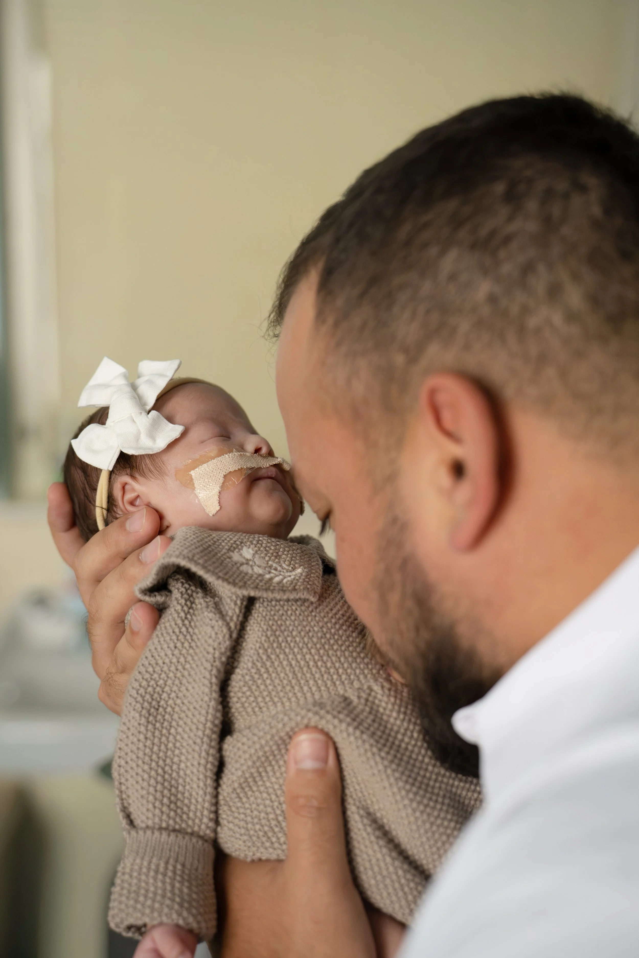 A man with a short haircut is holding a newborn baby girl with a large white bow in her hair, and a bandage over her nose, close to his face in a hospital room at Houston medical center.