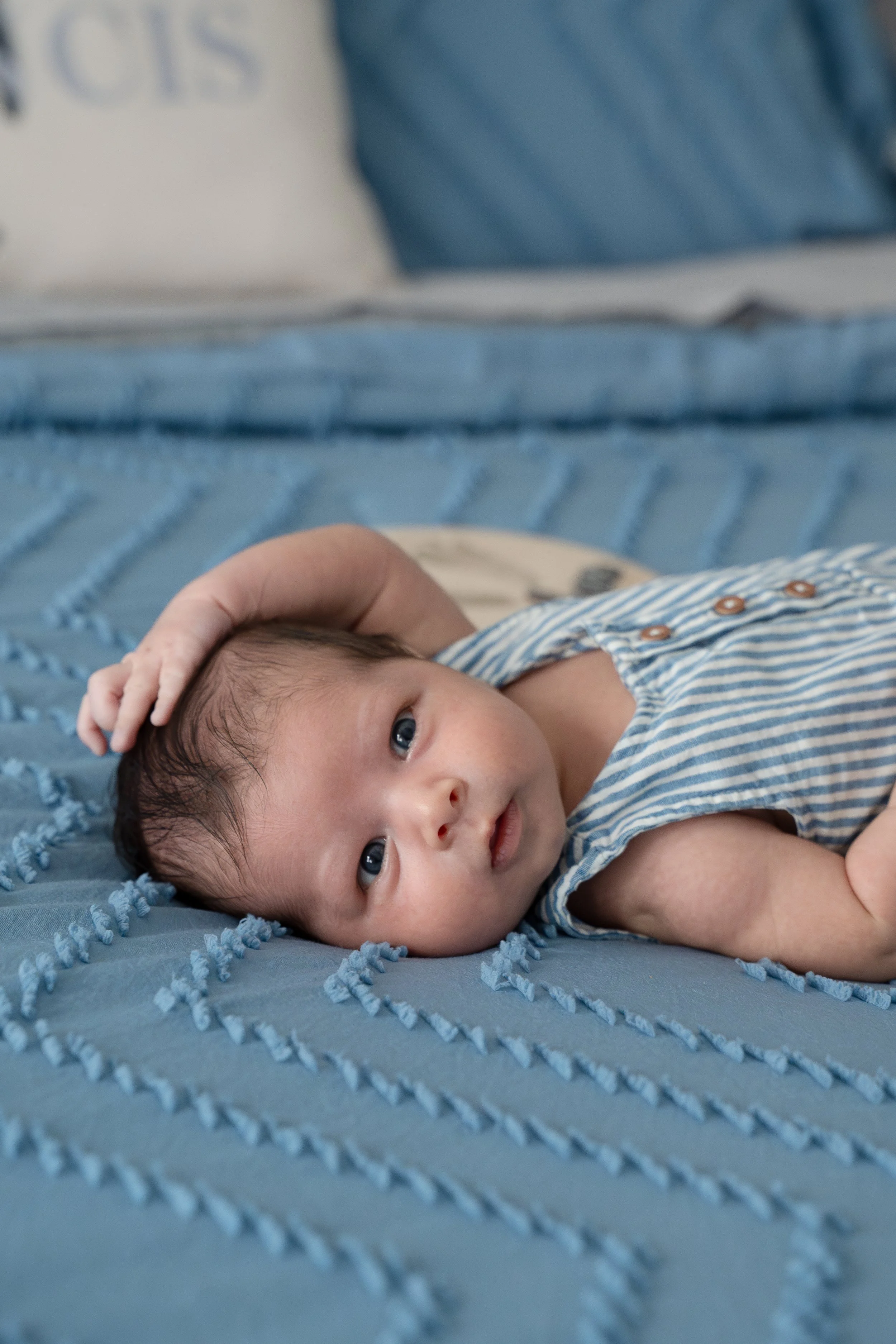 Close-up of a baby lying on a blue textured blanket, wearing a striped shirt, looking at the camera with wide eyes, in their The Woodlands home.