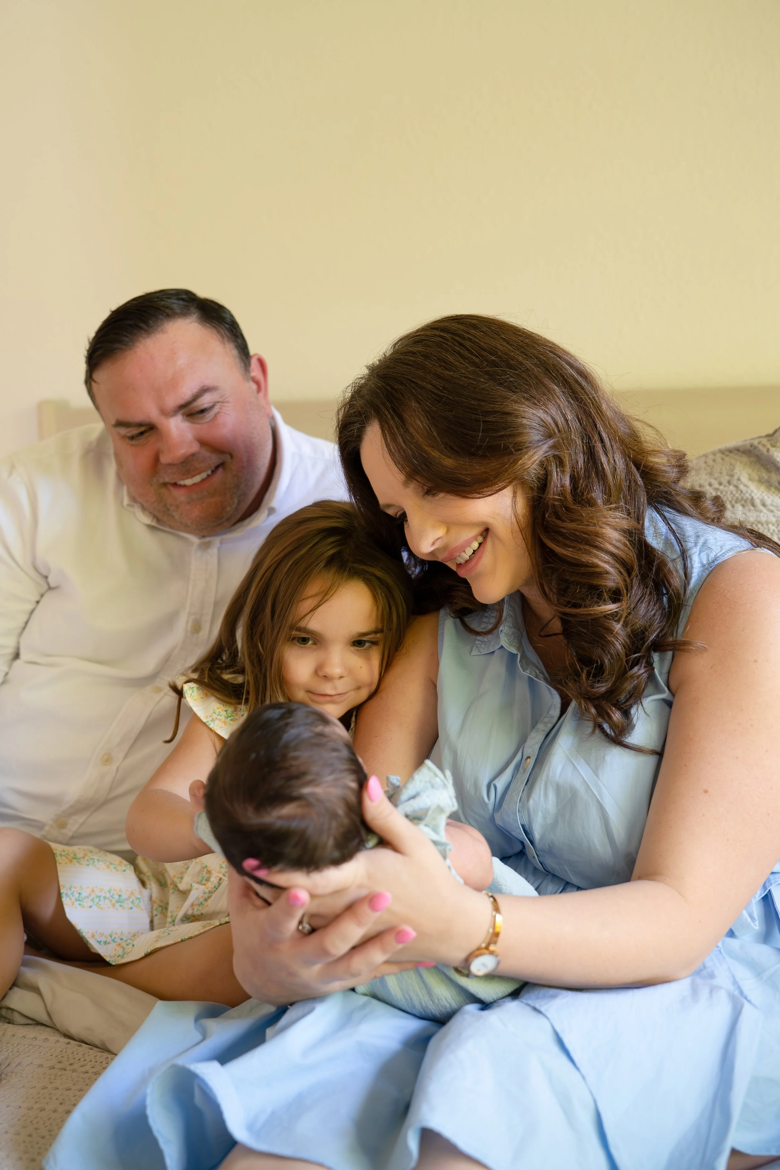 A family with two adults and two children gathered on a bed, looking at a baby, on the bed of their The Woodlands home.