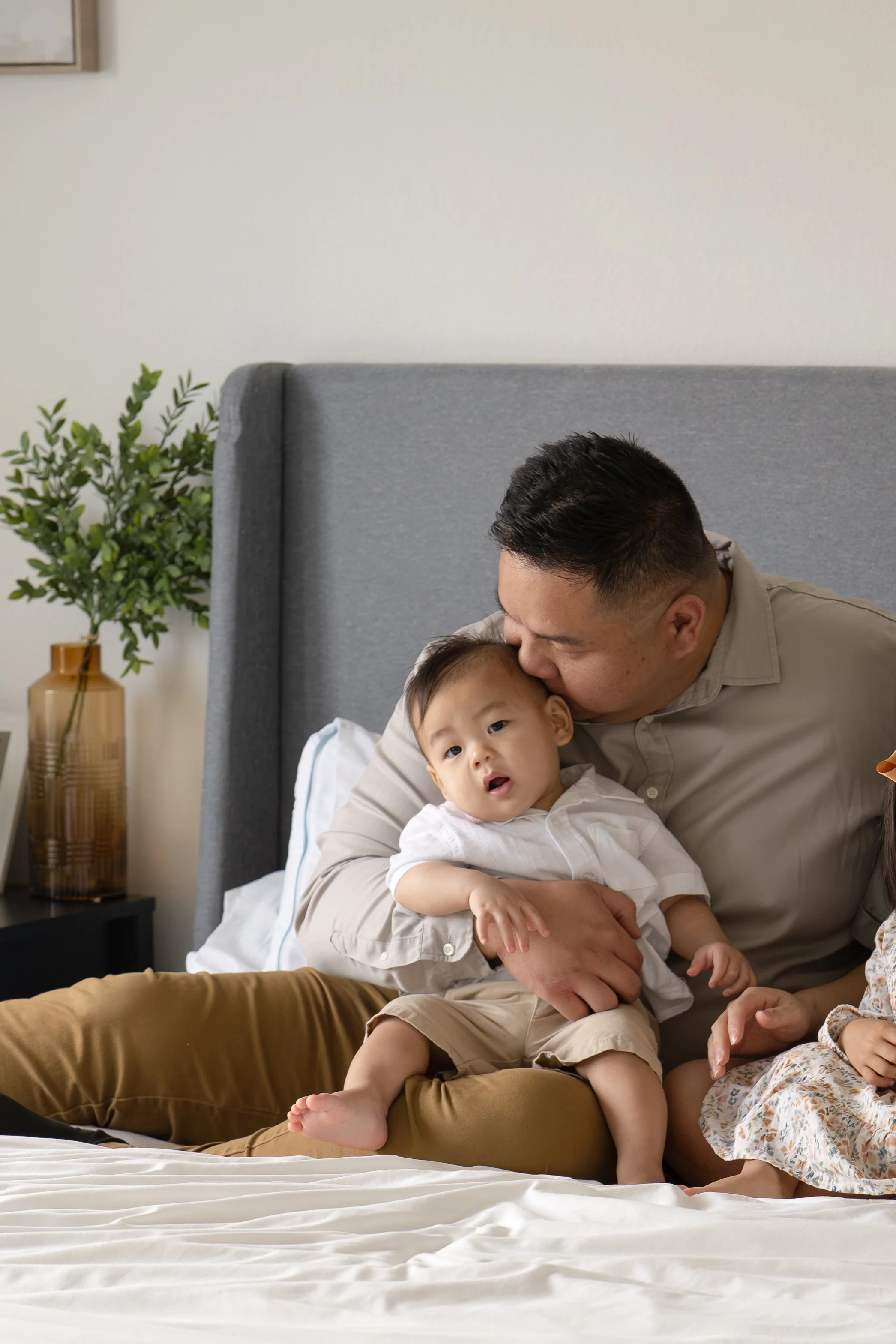A father and young son sitting on a bed, with the father kissing the son's head and the son looking at the camera, in a bedroom in Katy, TX home.
