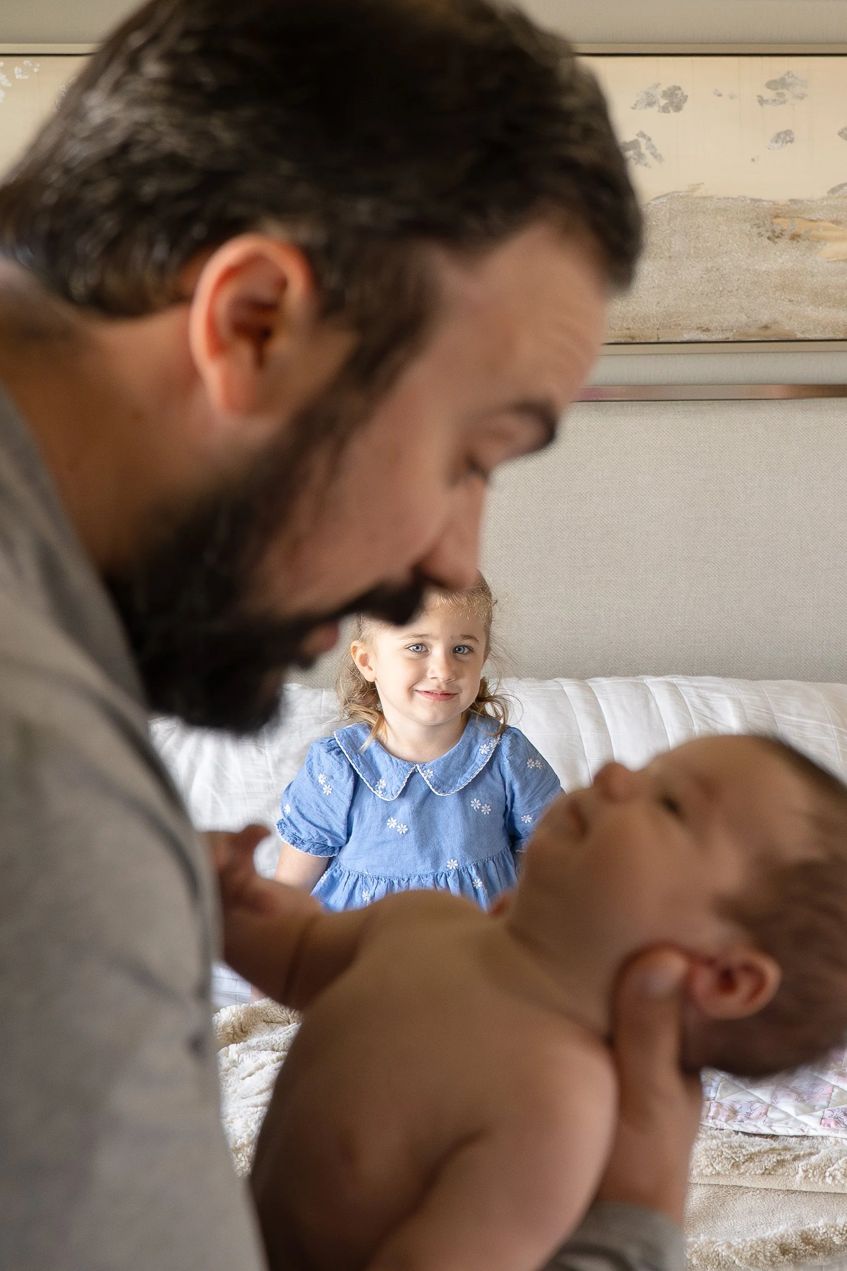 A man and a young girl are lying on a bed, with the man and baby in the foreground and the girl in the background, smiling and looking at the camera.  The Woodlands & Houston Heights In Home Newborn & Family photographer providing luxury artwork albu
