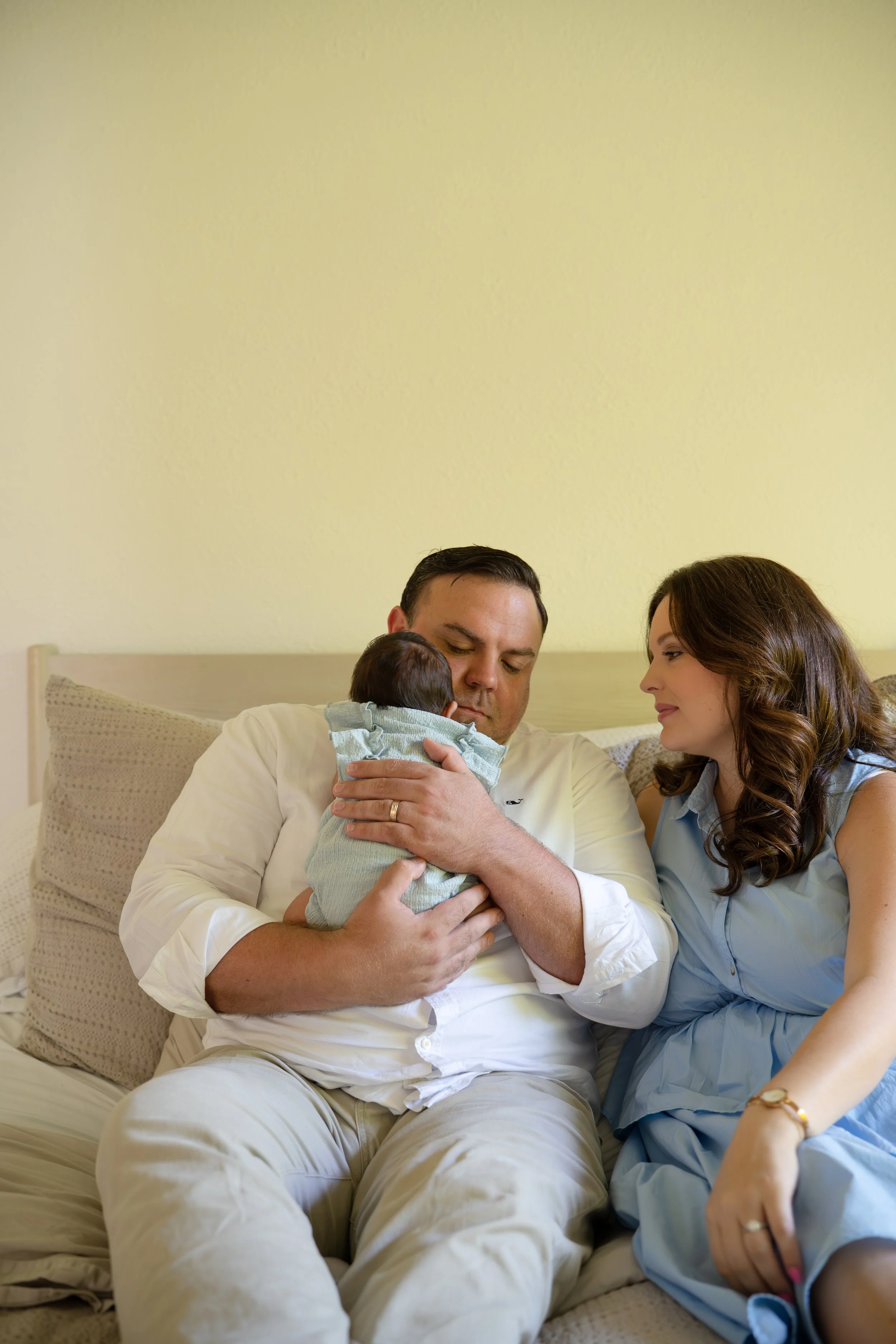 A family sitting on a bed: father holding a newborn baby, mother sitting beside him, all looking at the baby lovingly in their The Woodlands home.