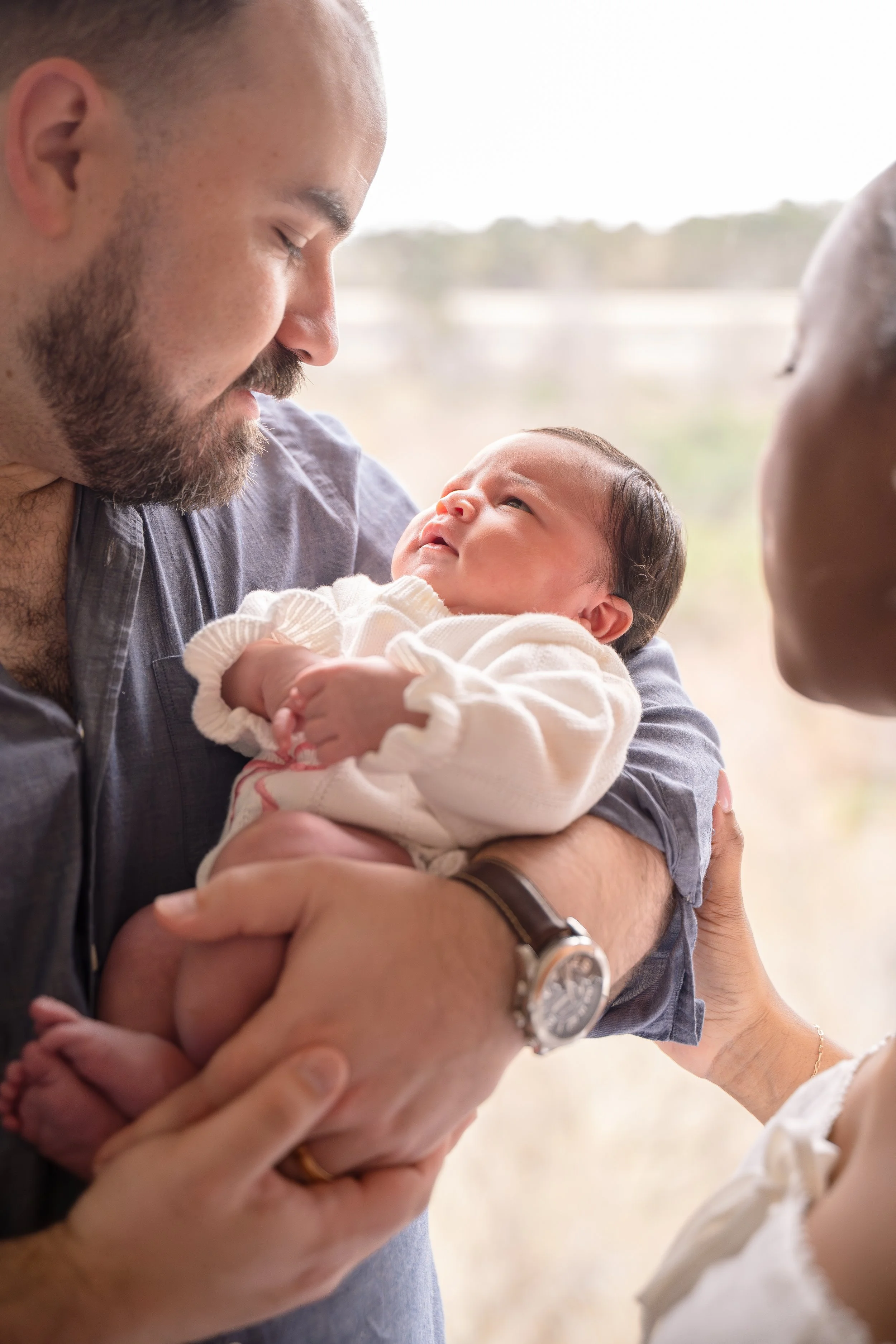 A man with a beard and a woman holding a newborn baby inside a room with a window overlooking a landscape. The man is smiling and looking at the baby, who is gazing up at him, in the Houston Ruby Postnatal. 