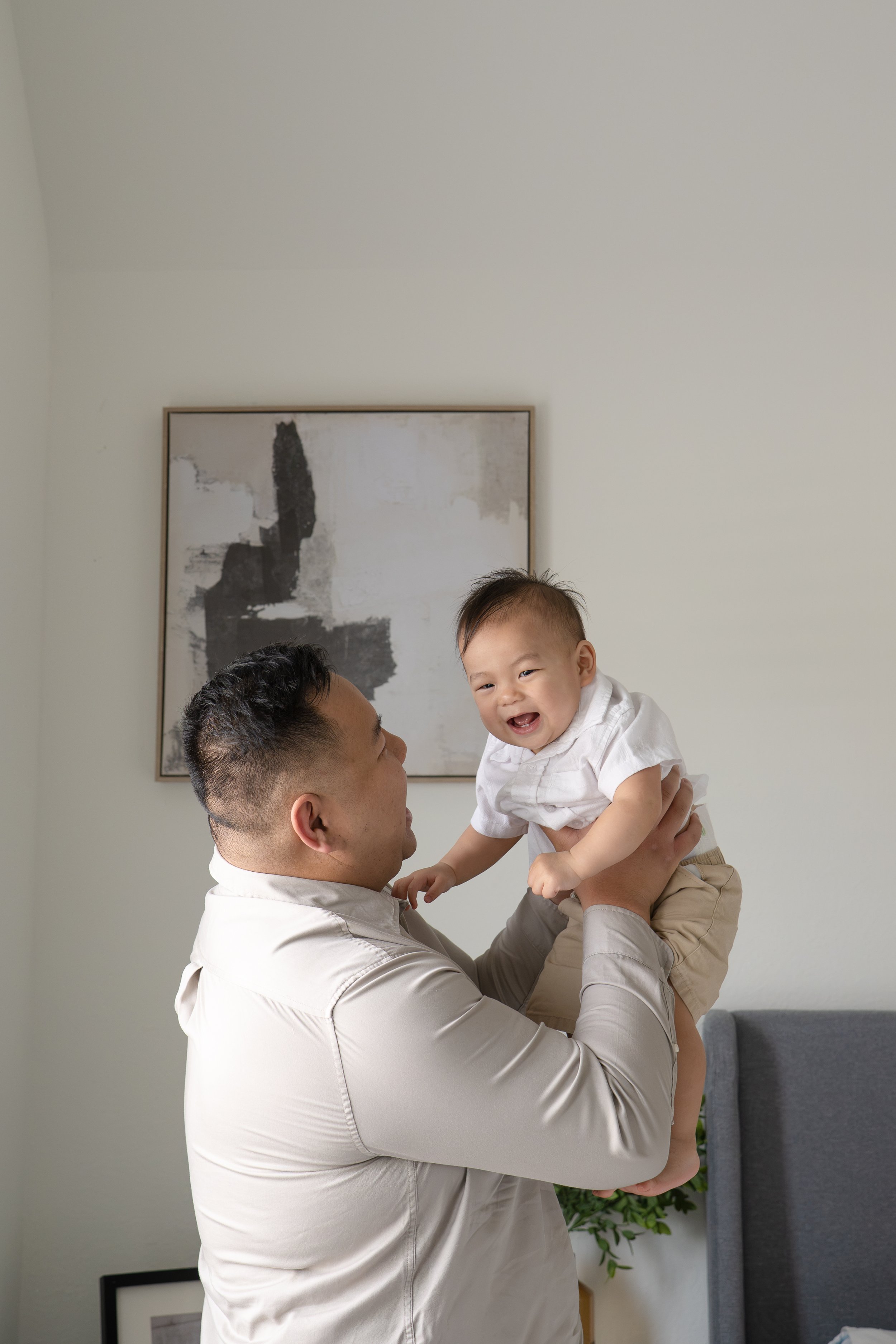 A man is lifting a smiling young boy dressed in white, indoors with a framed abstract black and white painting on the wall behind them. Playing with his dad at home in Katy, TX.