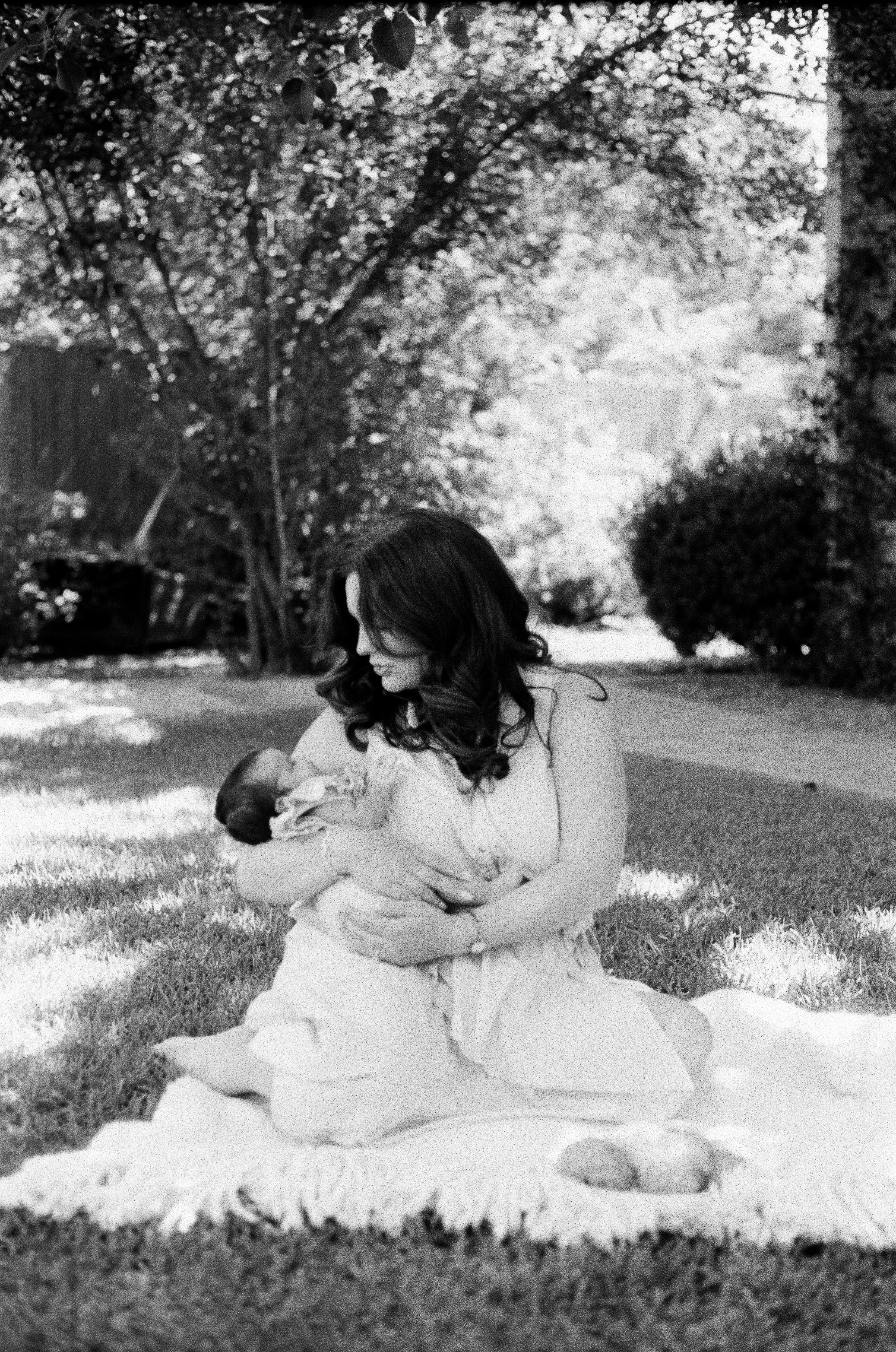 A woman sitting on the grass holding a baby in her arms in the backyard of her The Woodlands home with trees and bushes in the background.