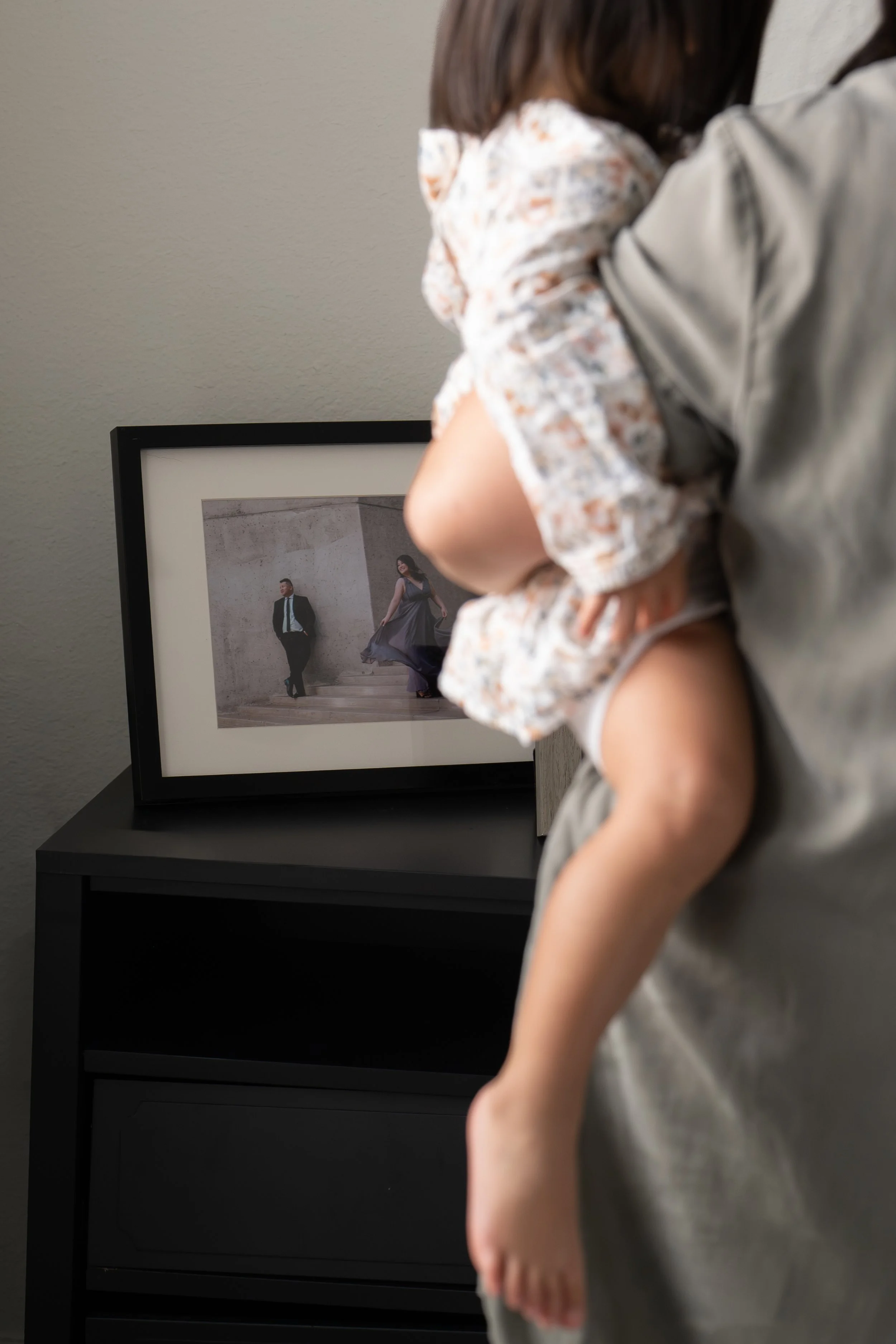 Child with curly hair wearing a floral blouse, standing next to an adult in gray, looking at a framed photograph of a man and woman dressed up and walking together, in Katy, TX at home.