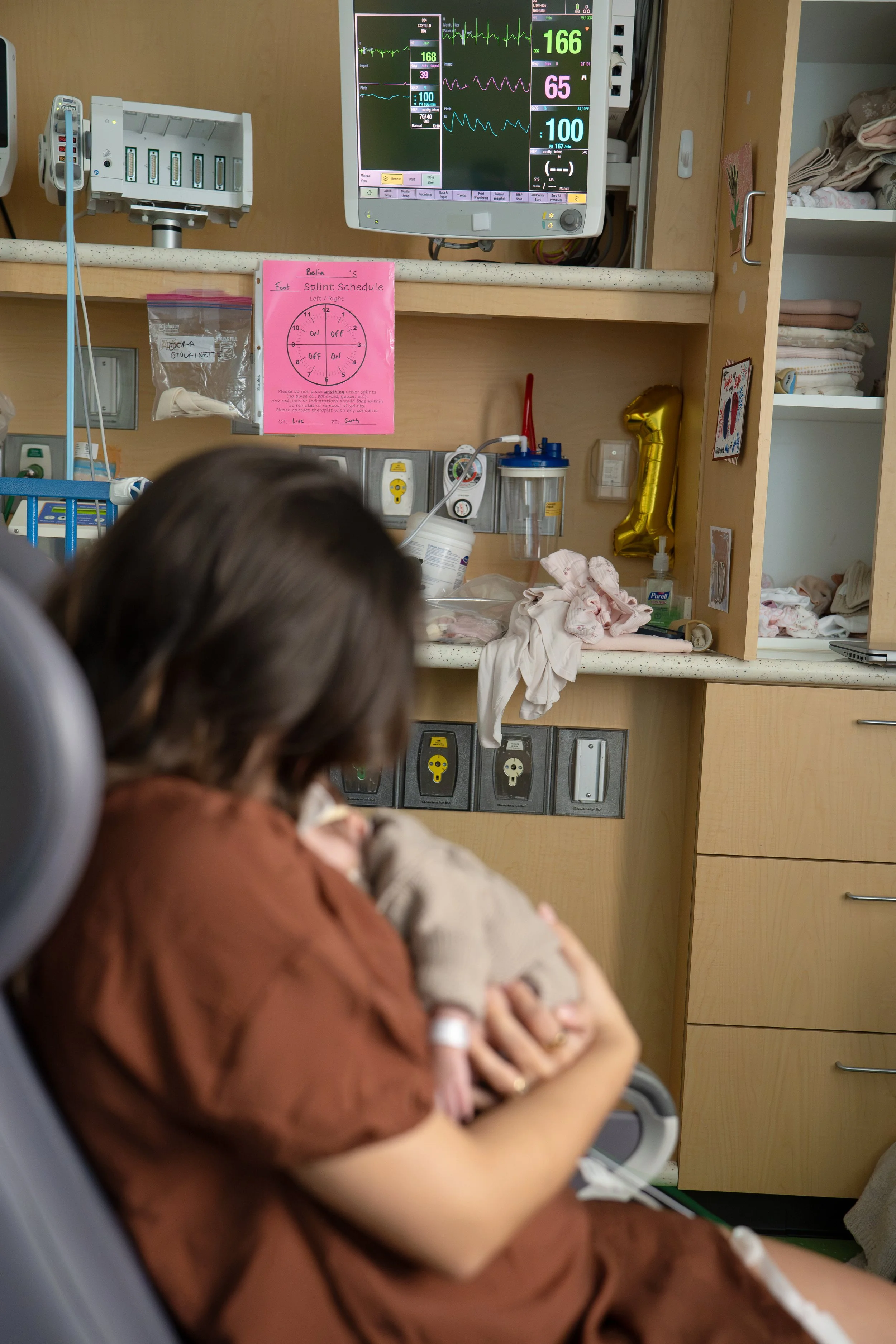 Close-up of a child in a hospital bed, holding a small stuffed animal. Medical equipment, including a monitor displaying vital signs and a shelf with folded blankets, at Houston medical center.