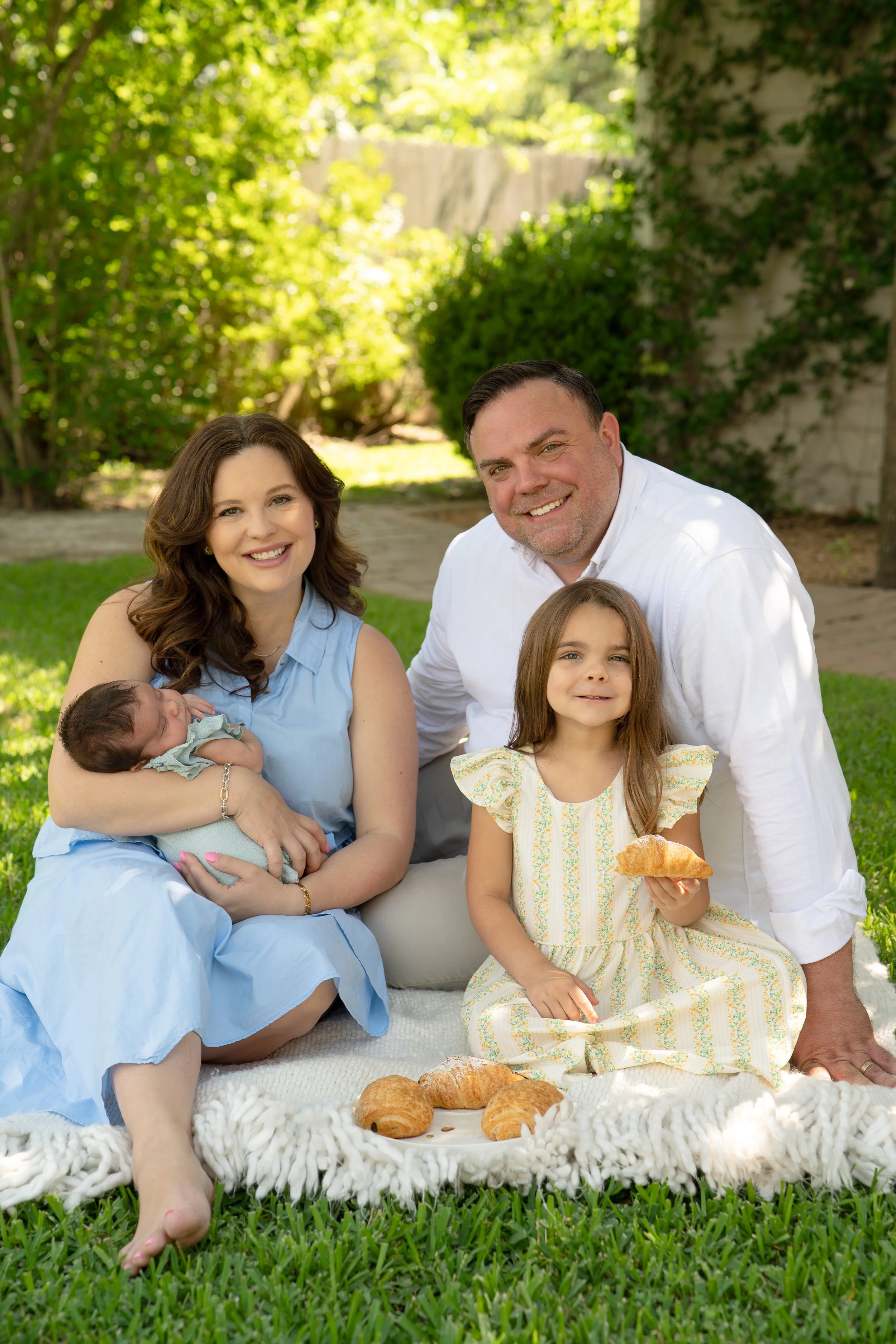 Family enjoying a picnic outdoors with a blanket, croissants, and a little girl holding a piece of bread in the backyard of their The Woodlands home.