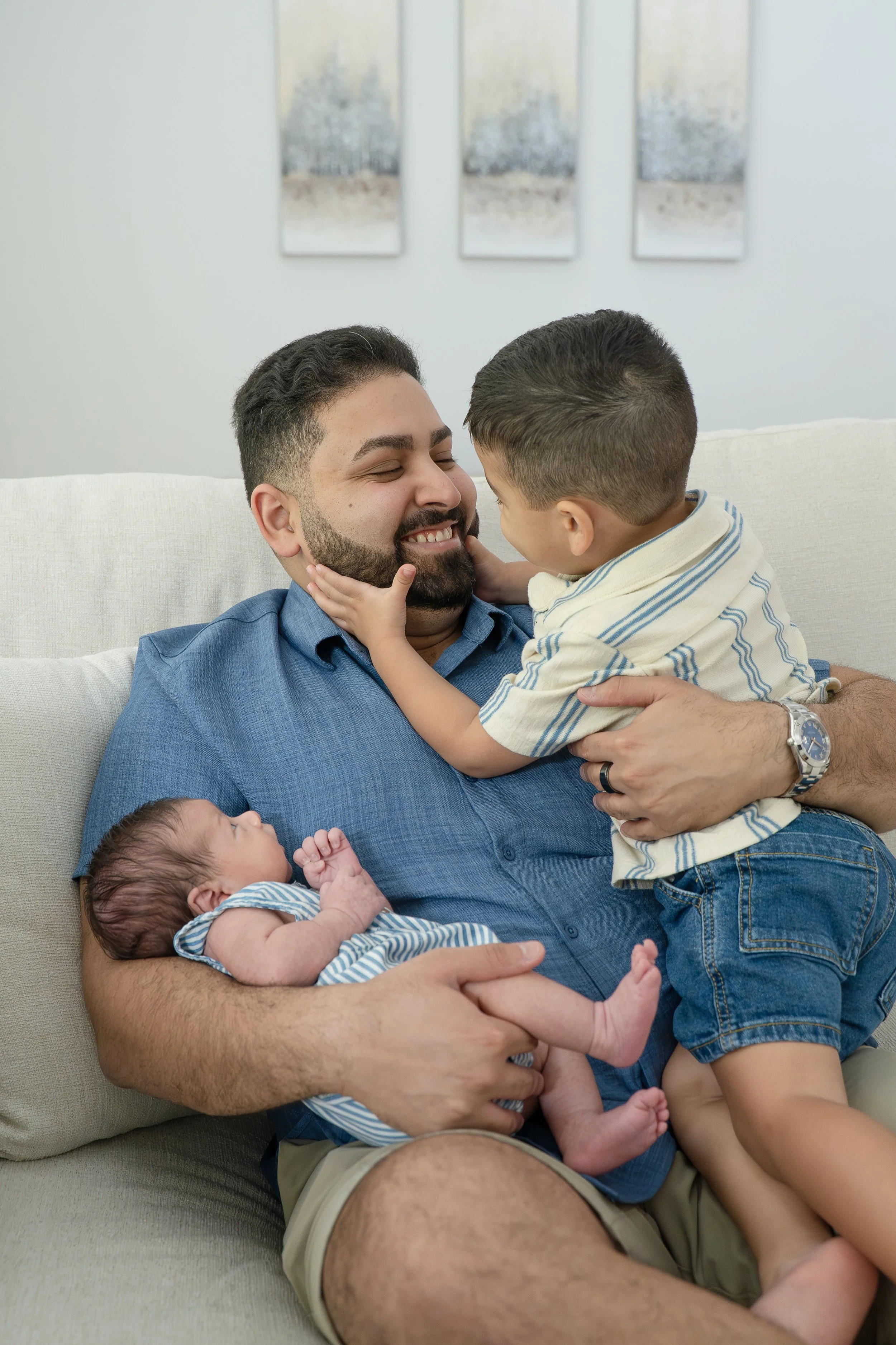 A man with a beard sitting on a beige couch, smiling as a young boy with short hair and a striped shirt kisses his nose, in their The Woodlands home.
