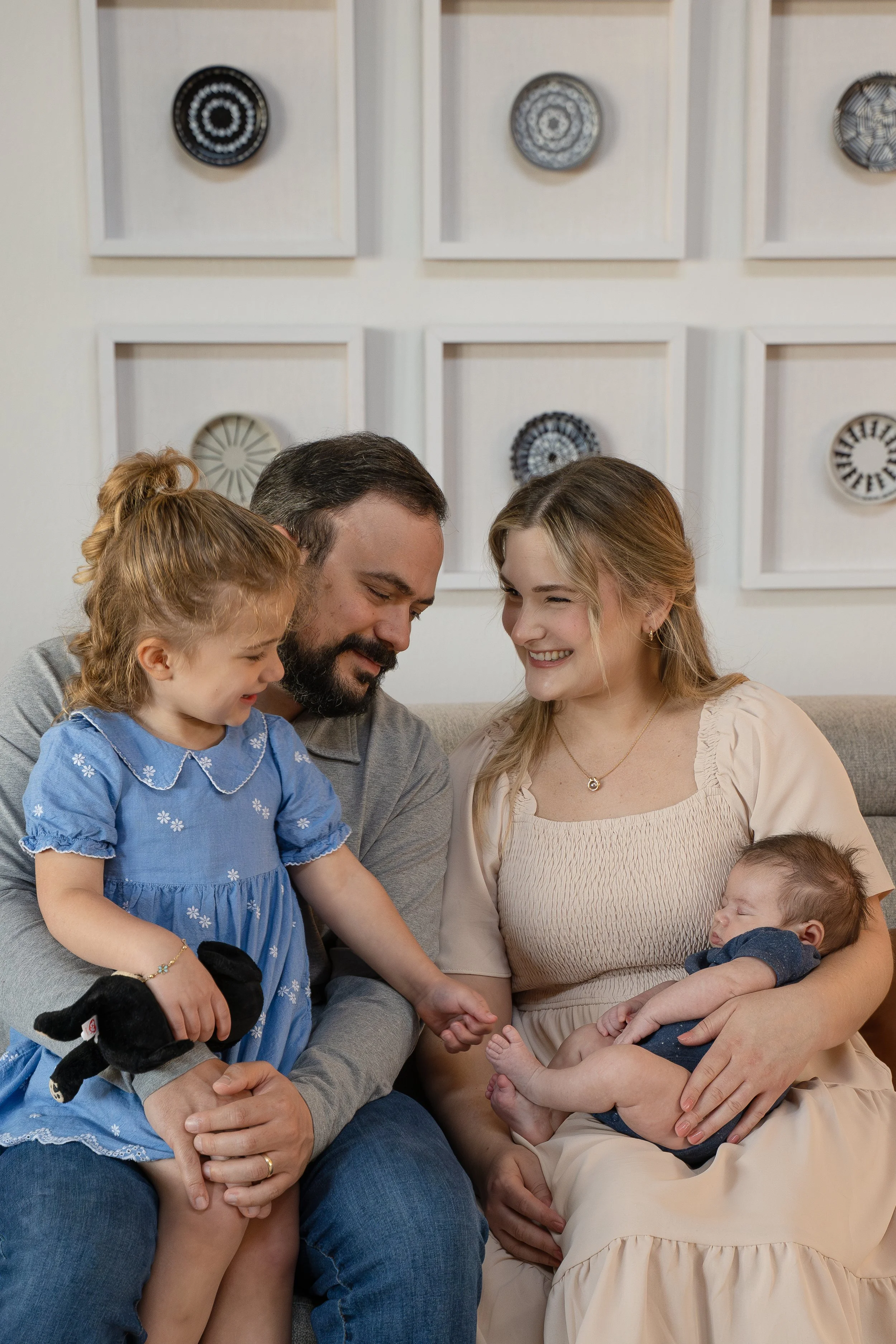 A family of five with two young children sitting on a couch, smiling and looking at a tablet device together. The background features wall art with circular designs.  The Woodlands & Houston Heights In Home Newborn & Family photographer providing lux