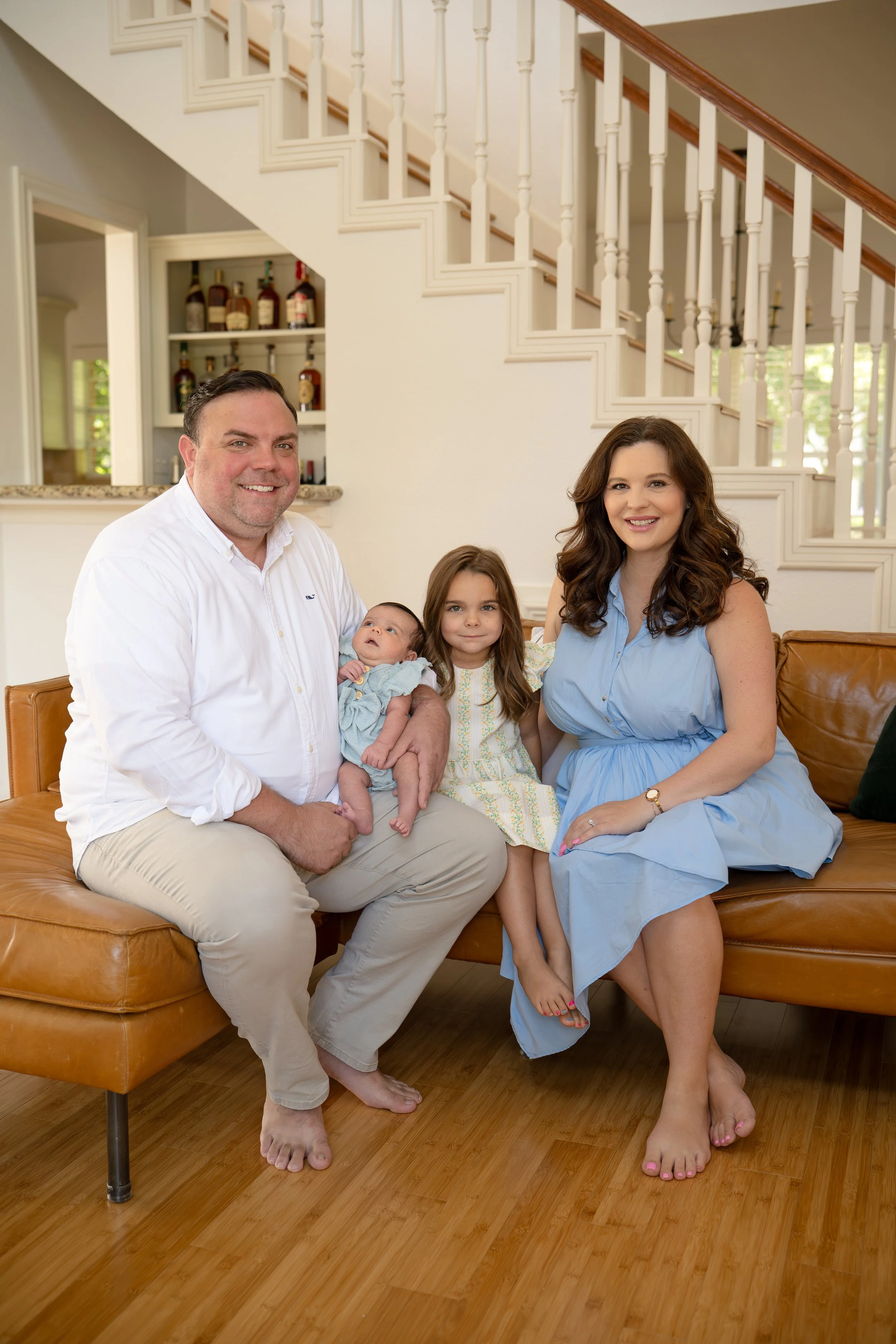 A smiling family sitting on a tan leather sofa in a bright living room with a staircase in the background in their new The Woodlands home. Dad is holding a newborn baby.