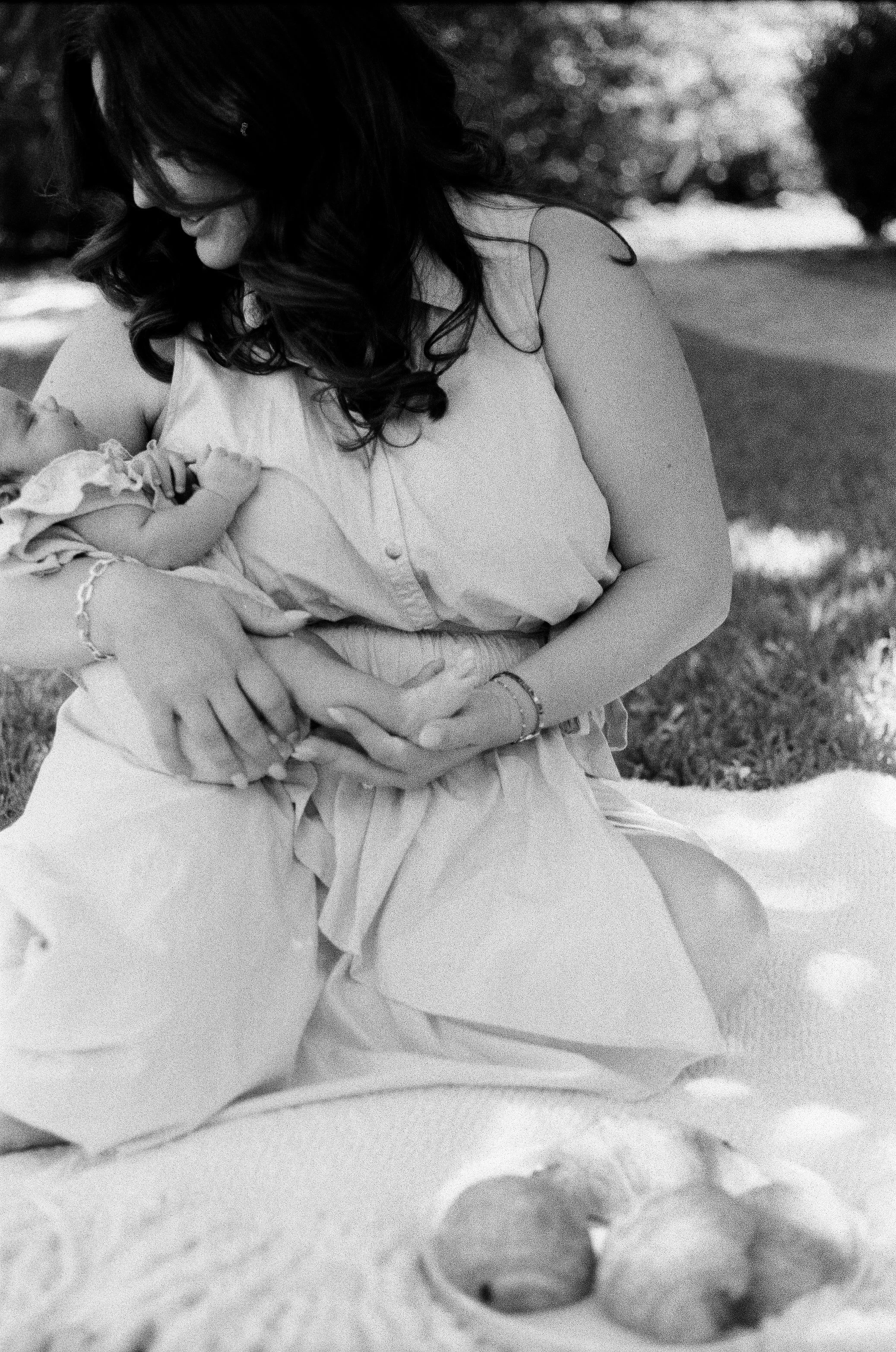 A woman with dark, curly hair and glasses is holding and interacting with a baby in their outdoor The Woodlands home, with trees and blurred greenery in the background.