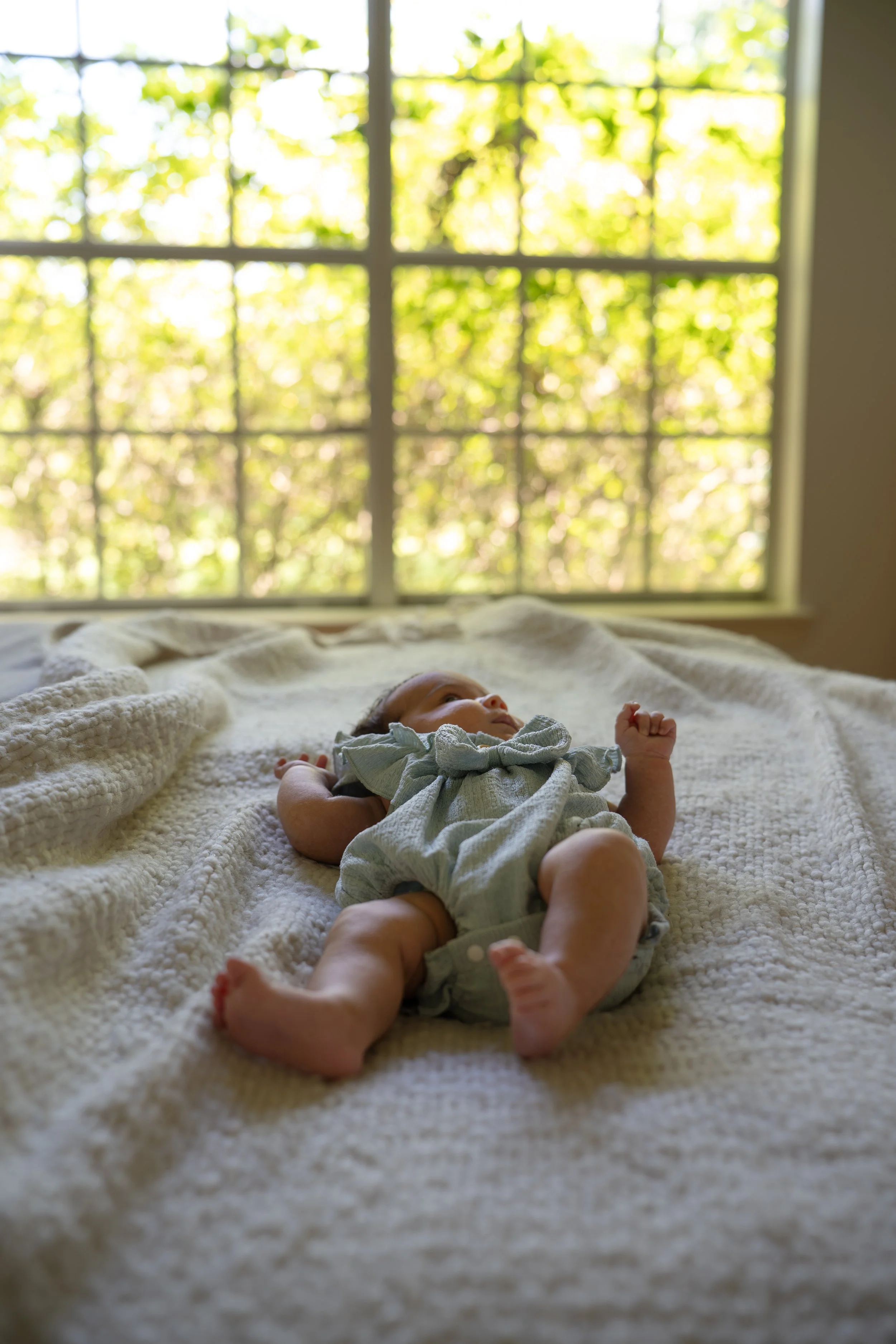 A baby lying on a bed covered with a white knit blanket, near a large window with sunlight and greenery outside. In their The Woodlands home.