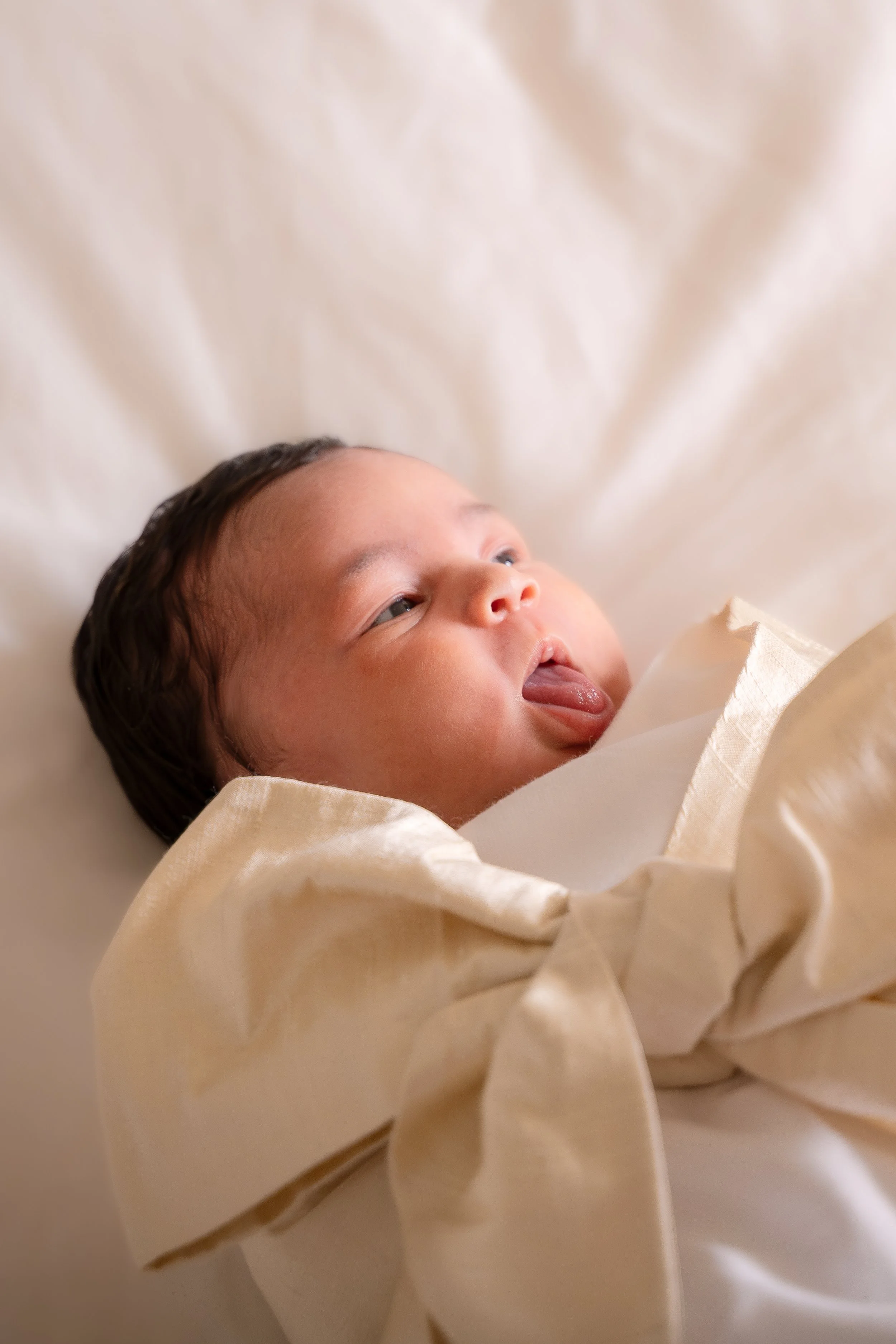 Close-up of a baby lying on a cream-colored blanket, with its face looking slightly upwards and tongue sticking out, dressed in a cream-colored outfit,in the Houston Ruby Postnatal. 