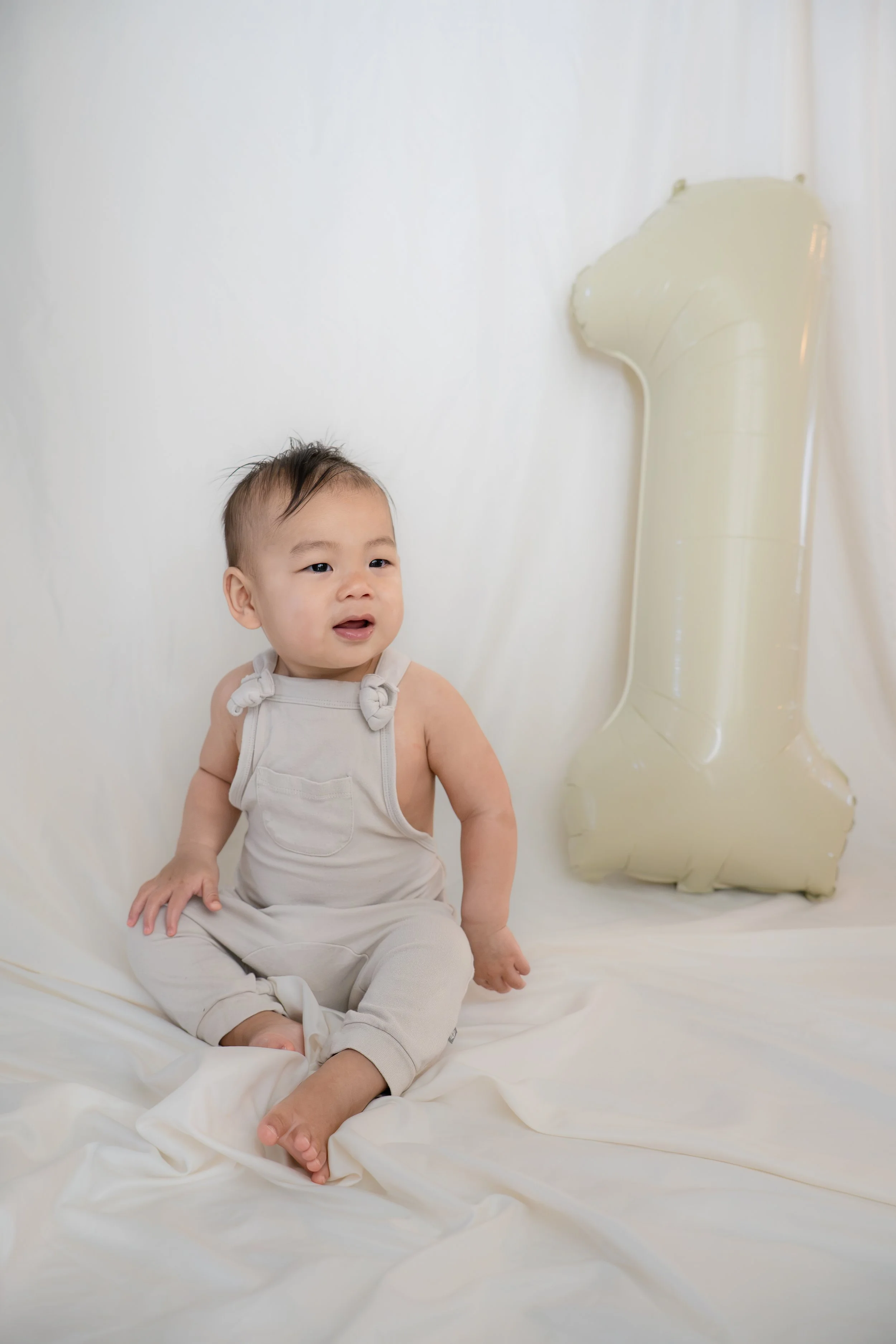A young child with short dark hair and light skin sitting on a white fabric surface, wearing a beige sleeveless outfit with bow details. Katy, TX toddler smiling with number 1.