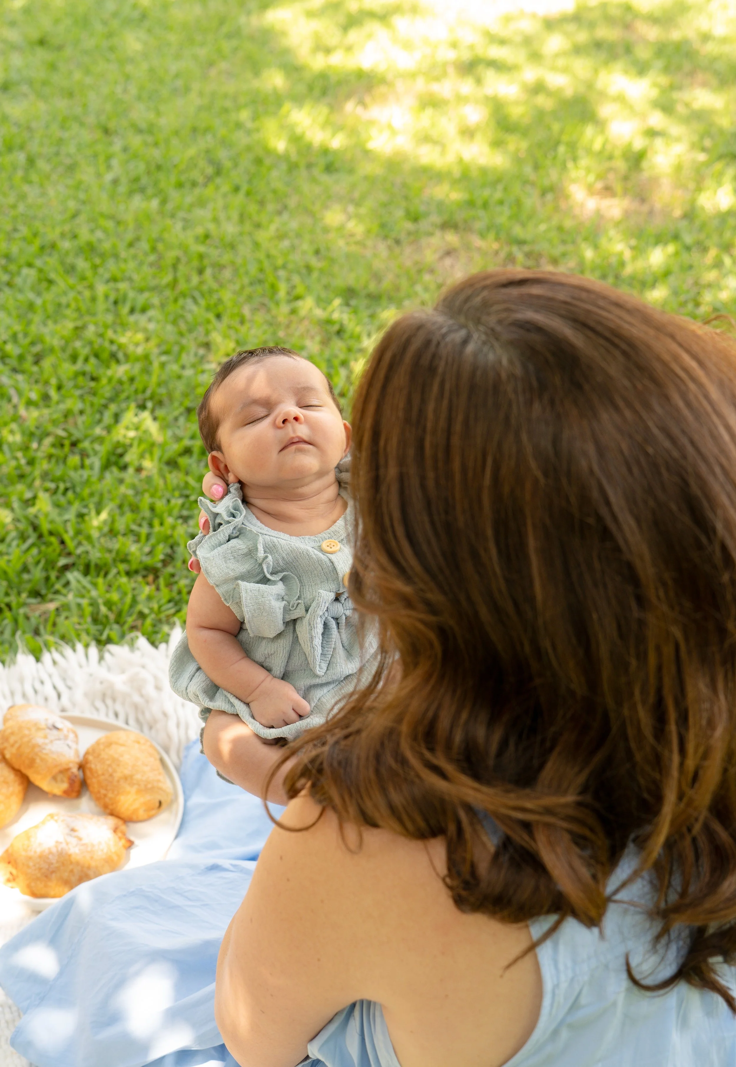A woman with long brown hair holding a sleeping baby with short dark hair outdoors on a grassy area, with a white blanket and a plate of croissants nearby, outdoor of their The Woodlands home.
